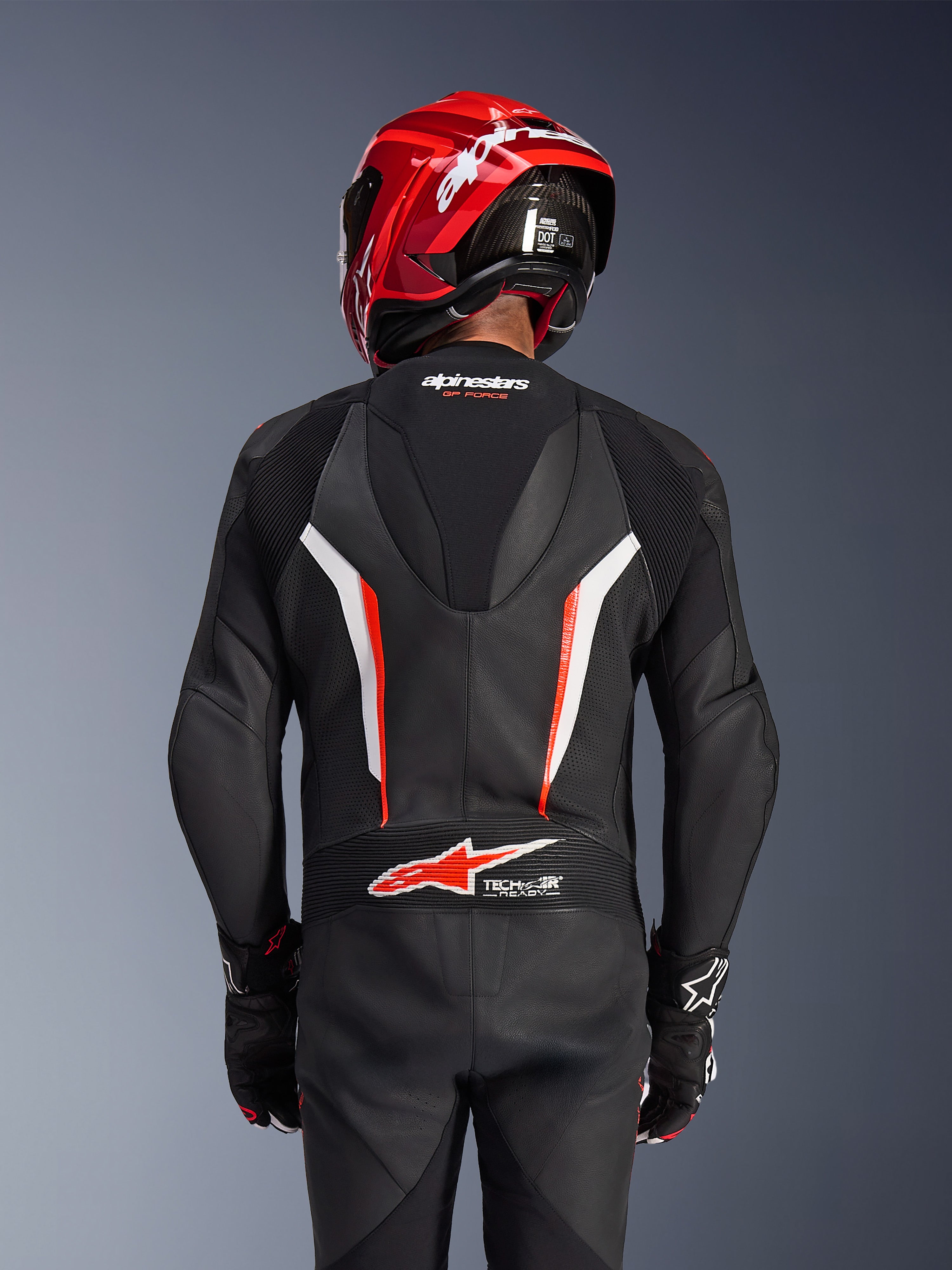 Combinaison en cuir Alpinestars GP Force V2 1 pièce, noir, blanc et rouge fluo, vue de dos d'un pilote portant un casque de course rouge et des gants, avec panneaux en cuir perforé, bosse aérodynamique avec logo Alpinestars et logo Tech-Air Ready dans le bas du dos, équipement de course moto professionnel.