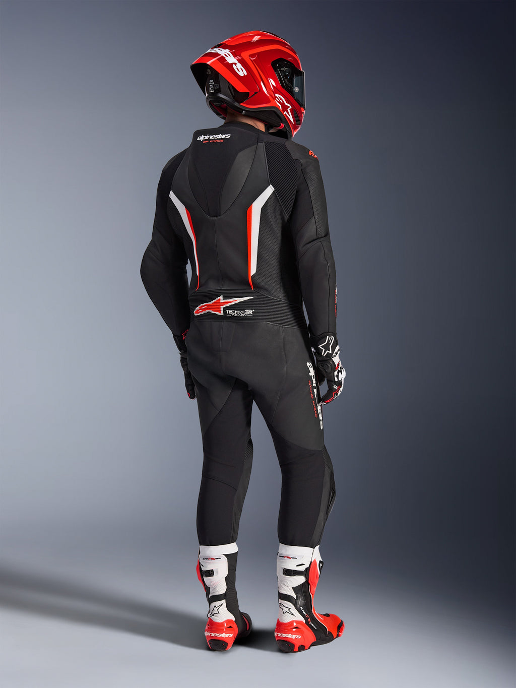 Pilote vu de dos portant la combinaison en cuir 1 pièce Alpinestars GP Force V2 en noir, blanc et rouge fluo, dotée d'une bosse aérodynamique et de panneaux perforés, avec casque rouge, gants de course et bottes de moto rouges et blanches, équipement professionnel de course sur circuit.