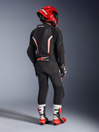 Pilote vu de dos portant la combinaison en cuir 1 pièce Alpinestars GP Force V2 en noir, blanc et rouge fluo, dotée d'une bosse aérodynamique et de panneaux perforés, avec casque rouge, gants de course et bottes de moto rouges et blanches, équipement professionnel de course sur circuit.