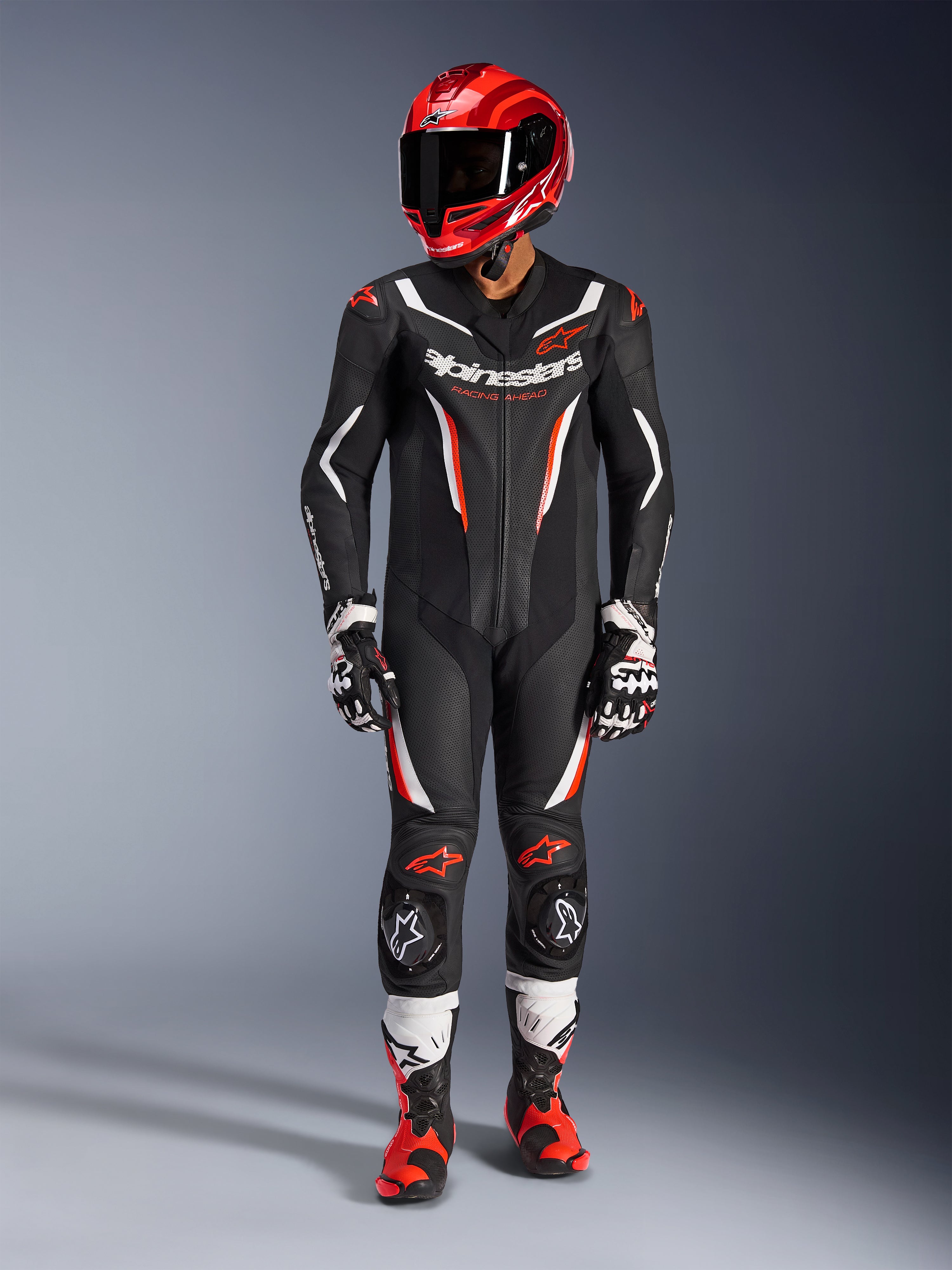 Combinaison en cuir Alpinestars GP Force V2 1 pièce, noir, blanc et rouge fluo, portée avec un casque de course rouge, des gants noirs et blancs et des bottes rouges et noires, dotée de panneaux en cuir perforé et du logo Alpinestars, équipement professionnel de course de moto