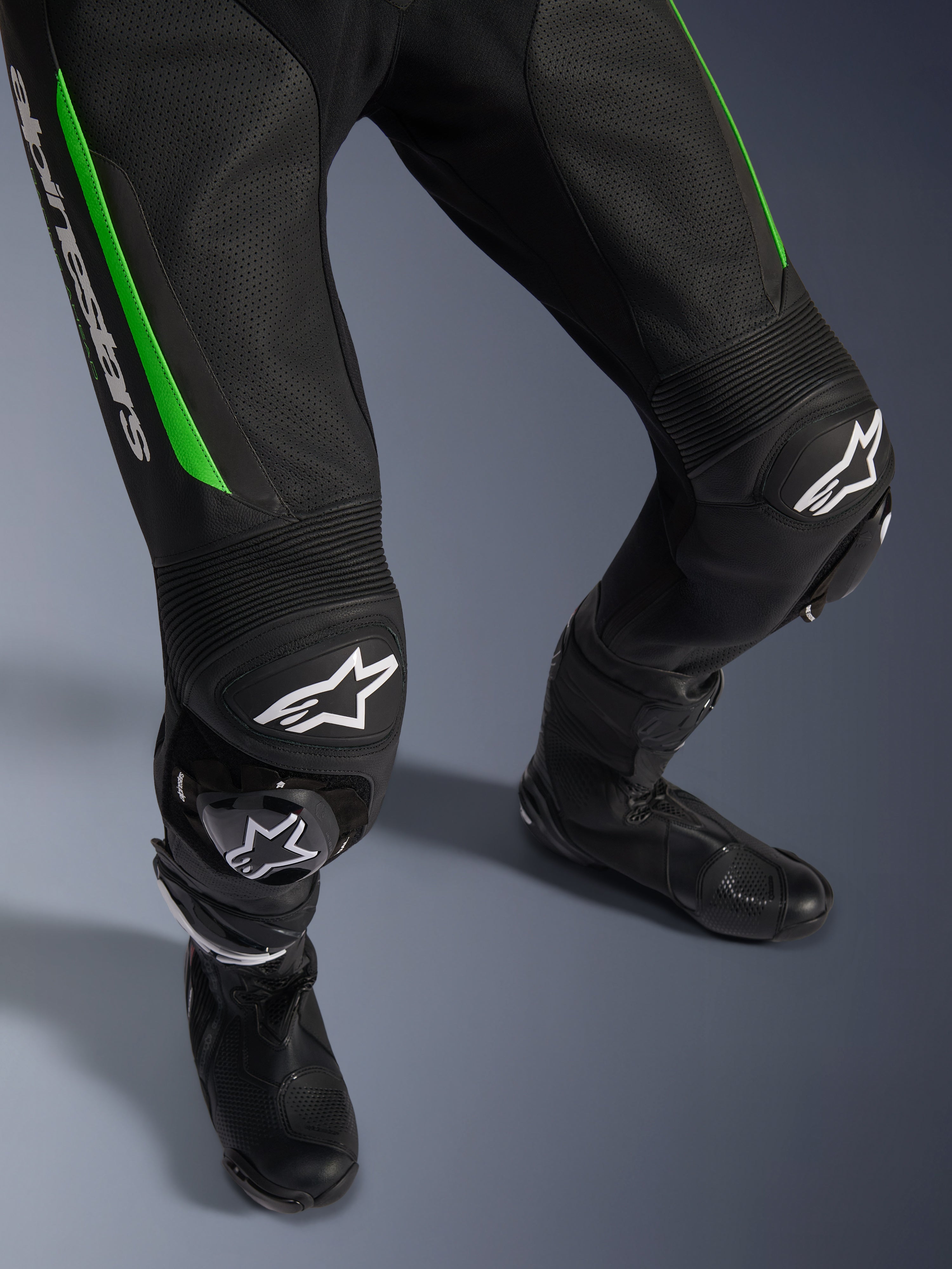 Alpinestars GP Force V2 Leather Suit - 1 pièce, gros plan sur les jambes en noir et vert, présentant une construction en cuir perforé, des panneaux accordéon aux genoux, le marquage Alpinestars blanc et des sliders de genoux intégrés, portée avec des bottes de course noires