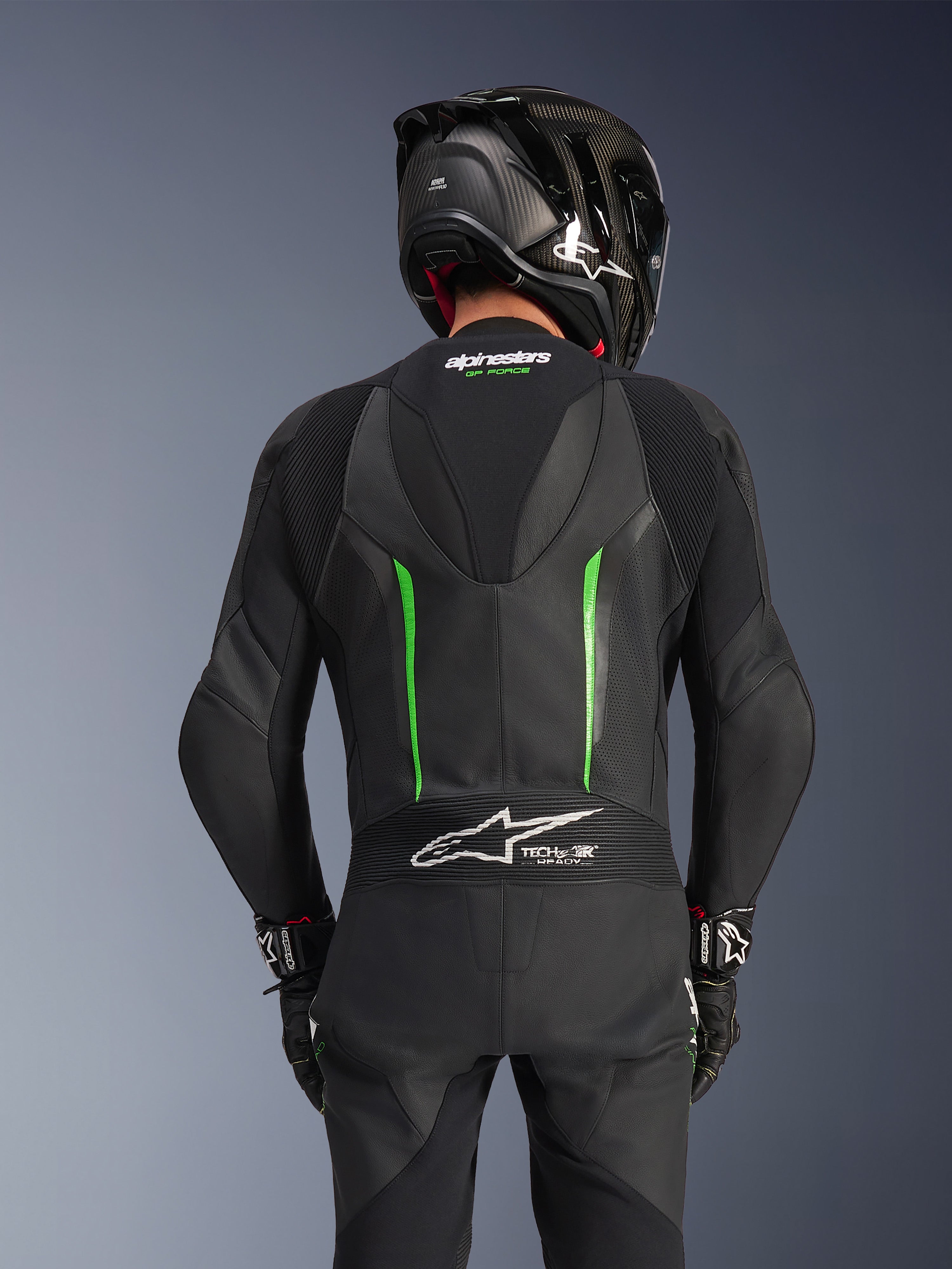 Vue arrière d'un pilote portant la combinaison en cuir Alpinestars GP Force V2 en Noir Vert, dotée d'une bosse aérodynamique, du marquage Tech-Air Ready sur le panneau accordéon au bas du dos et d'une construction en cuir perforé, associée à un casque de course noir en fibre de carbone.