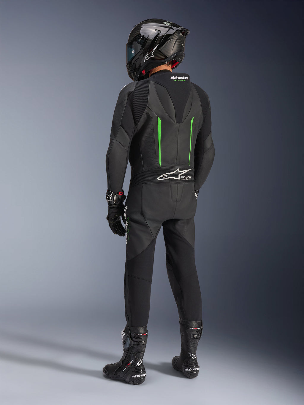 Pilote de dos portant une combinaison en cuir Alpinestars GP Force V2, 1 pièce, noir avec accents verts, bosse aérodynamique, avec un casque noir en fibre de carbone, des bottes de course et des gants noirs, équipement de course moto haute performance