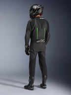 Pilote de dos portant une combinaison en cuir Alpinestars GP Force V2, 1 pièce, noir avec accents verts, bosse aérodynamique, avec un casque noir en fibre de carbone, des bottes de course et des gants noirs, équipement de course moto haute performance