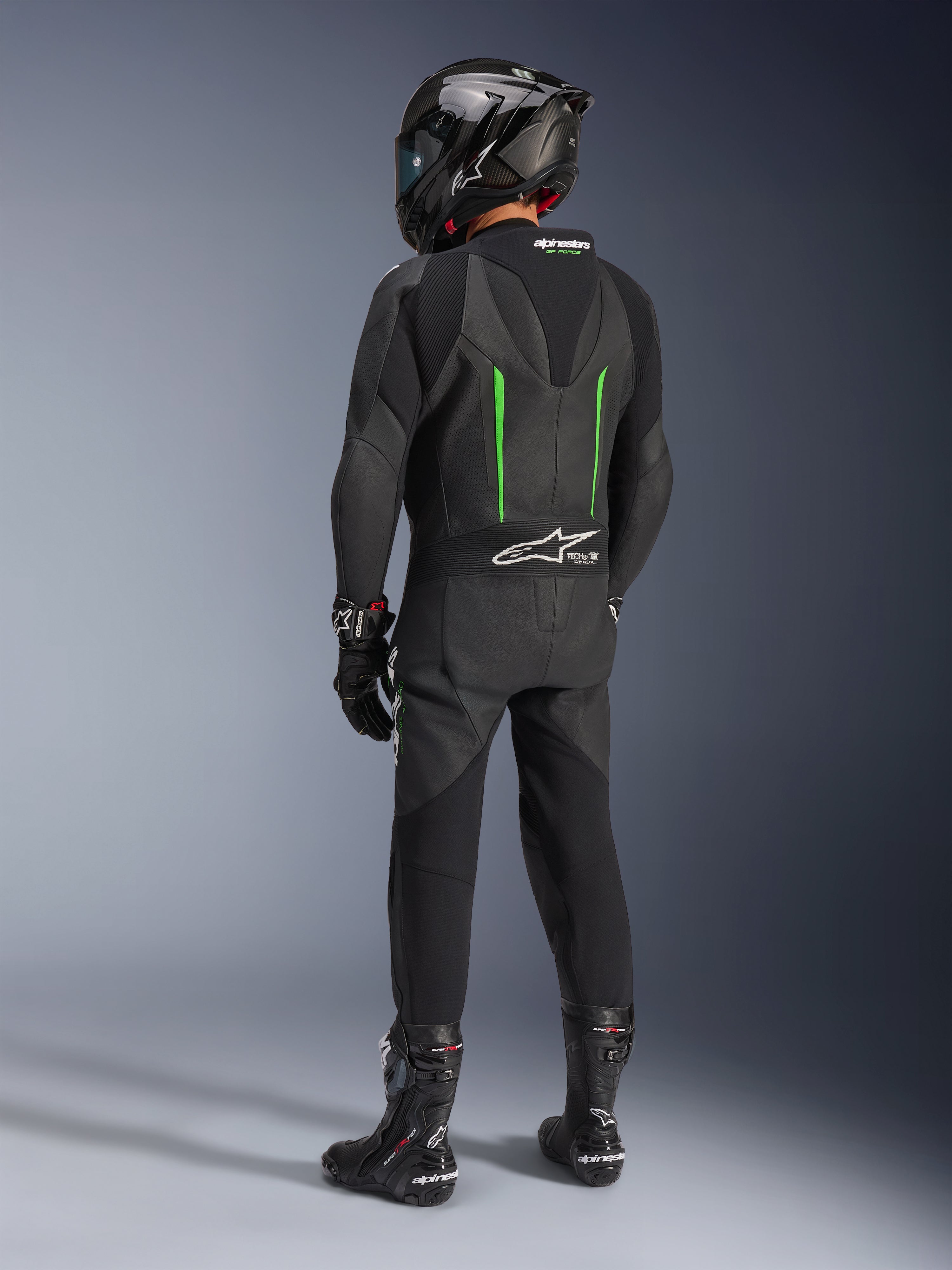 Pilote de dos portant une combinaison en cuir Alpinestars GP Force V2, 1 pièce, noir avec accents verts, bosse aérodynamique, avec un casque noir en fibre de carbone, des bottes de course et des gants noirs, équipement de course moto haute performance