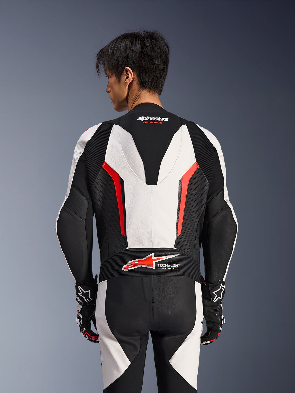 Combinaison en cuir Alpinestars GP Force V2 1 pièce, blanc, noir et rouge fluo, vue de dos sur un modèle masculin, avec bosse aérodynamique, panneaux en cuir perforé et logo Tech-Air Ready dans le bas du dos, équipement de course professionnel avec gants noirs et blancs