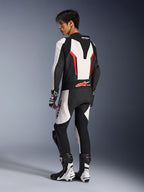 Pilote de moto portant la combinaison en cuir Alpinestars GP Force V2 - 1 pièce en blanc, noir et rouge fluo, vue arrière latérale montrant la bosse aérodynamique et les panneaux en cuir perforé, associée à des bottes et des gants de course assortis, conçue pour la piste et la performance.
