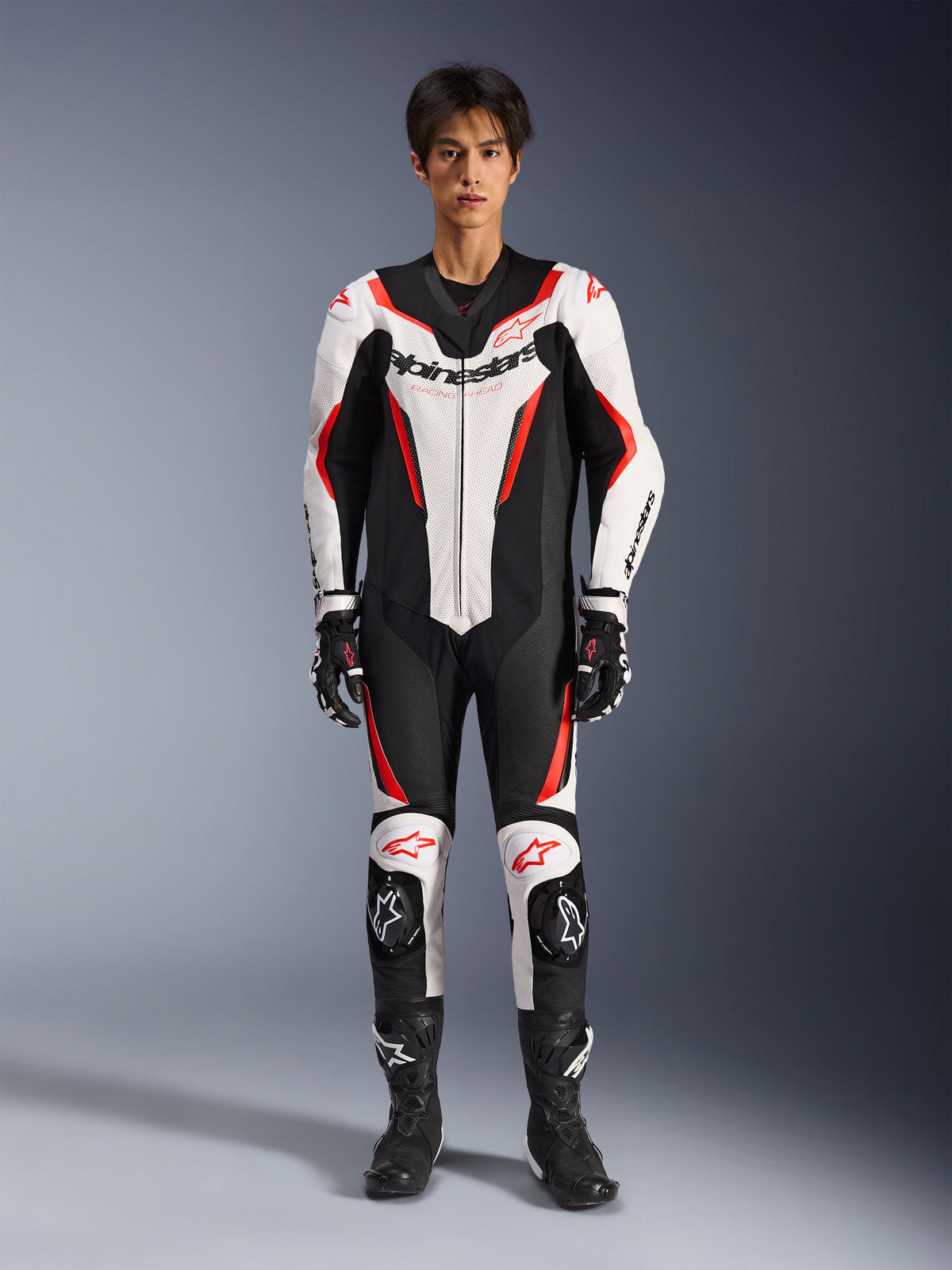 Combinaison en cuir Alpinestars GP Force V2 - 1 pièce, blanc, noir et rouge fluo, combinaison de moto de course monopièce, vue de face sur modèle, avec panneaux perforés et accents rouges, logo Alpinestars imposant sur la poitrine, assortie avec des bottes et des gants de course professionnels