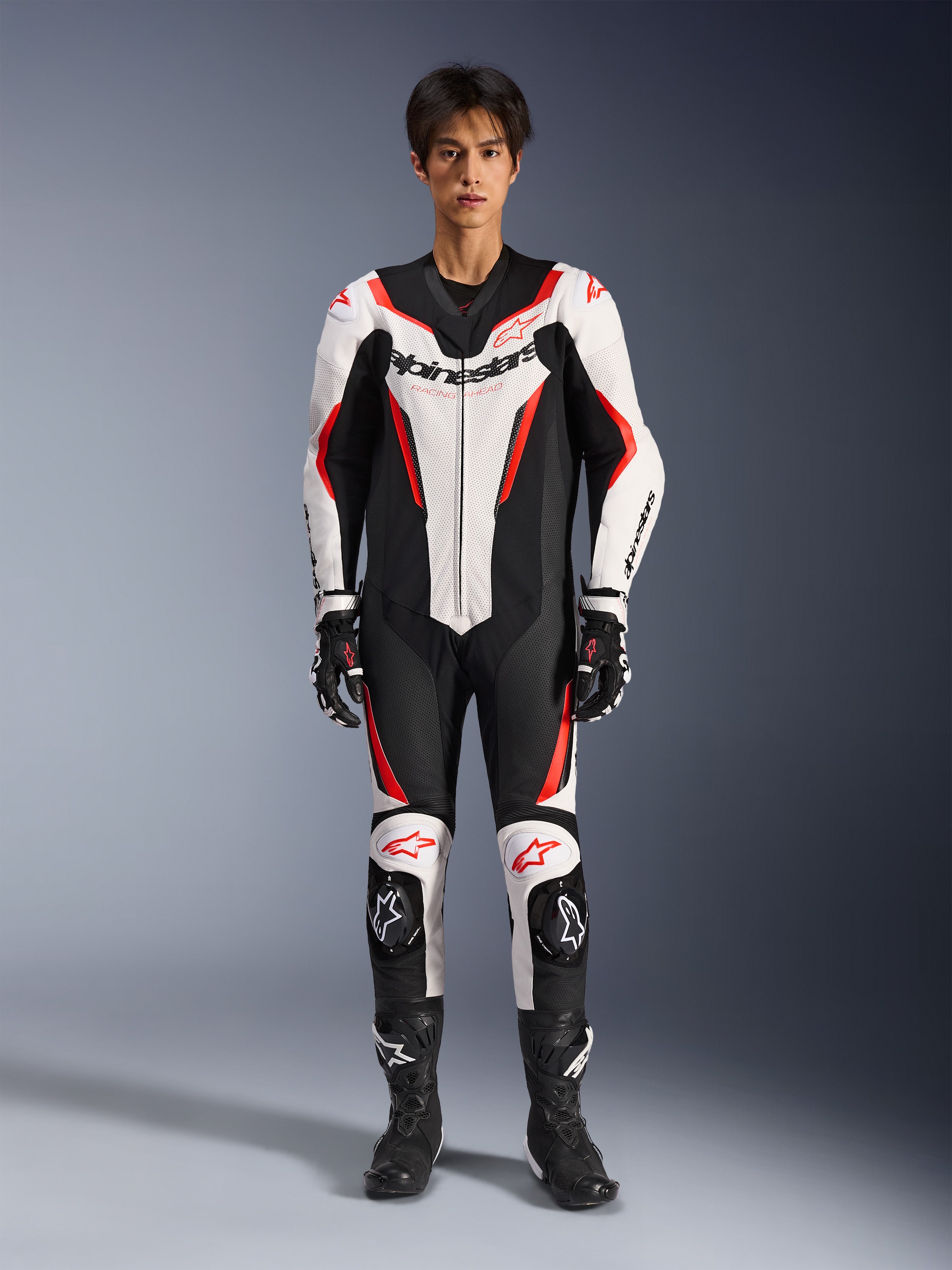 Combinaison en cuir Alpinestars GP Force V2 - 1 pièce, blanc, noir et rouge fluo, combinaison de course moto une pièce, portée par un mannequin avec gants et bottes de course assortis, logo Alpinestars distinctif, panneaux en cuir perforé et design sportif aérodynamique