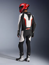 Combinaison en cuir Alpinestars GP Force V2 - 1 pièce, Blanc Noir Rouge Fluo, vue arrière d'un pilote portant un équipement de course complet, incluant un casque rouge, des bottes et des gants noirs et blancs, mettant en évidence la bosse aérodynamique et les panneaux extensibles ergonomiques.