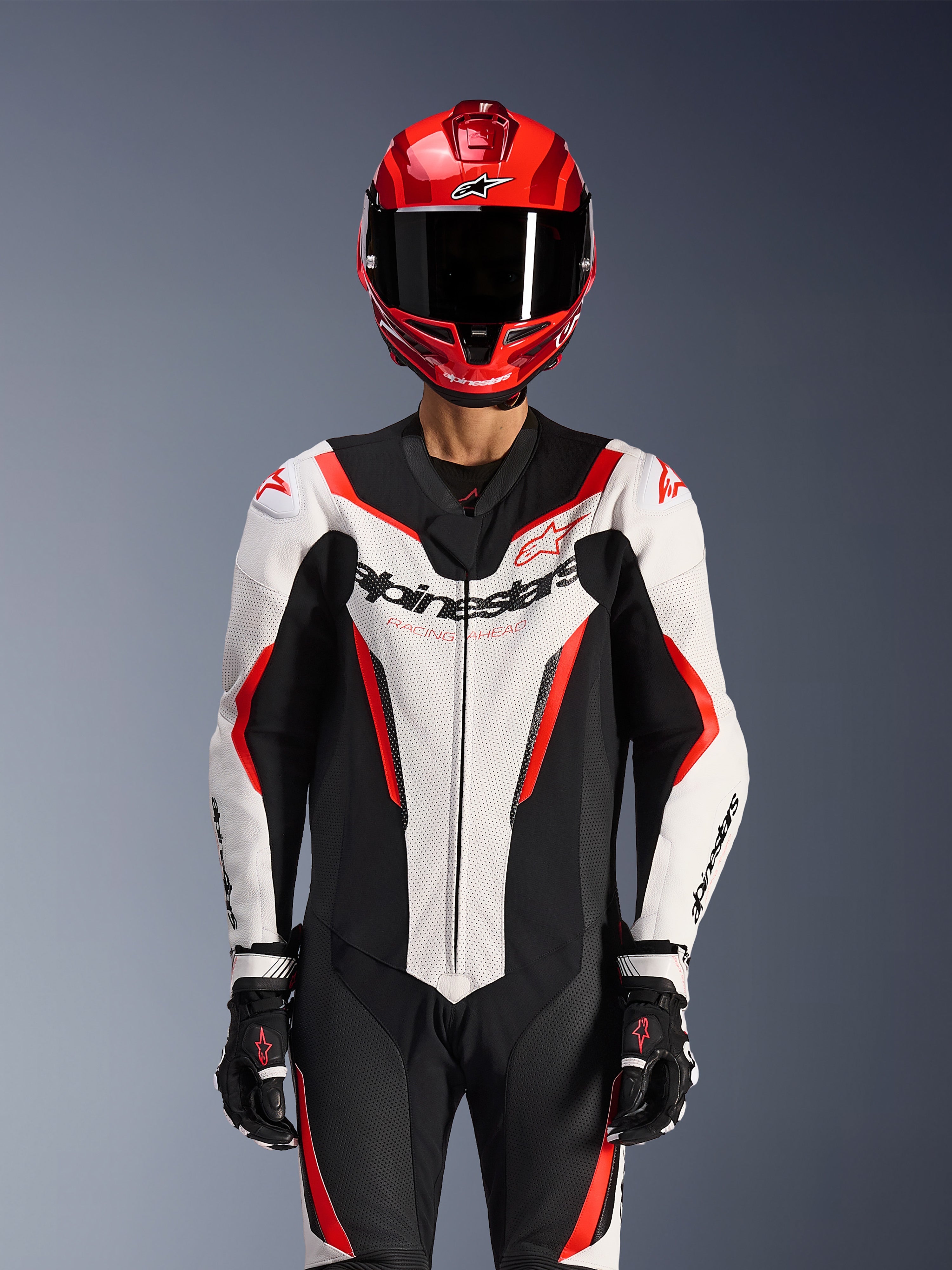 Combinaison en cuir Alpinestars GP Force V2 - 1 pièce, blanc, noir et rouge fluo, portée avec un casque intégral rouge et des gants de course noirs, panneaux en cuir perforé et logo Alpinestars sur la poitrine, équipement de performance professionnel pour la course moto
