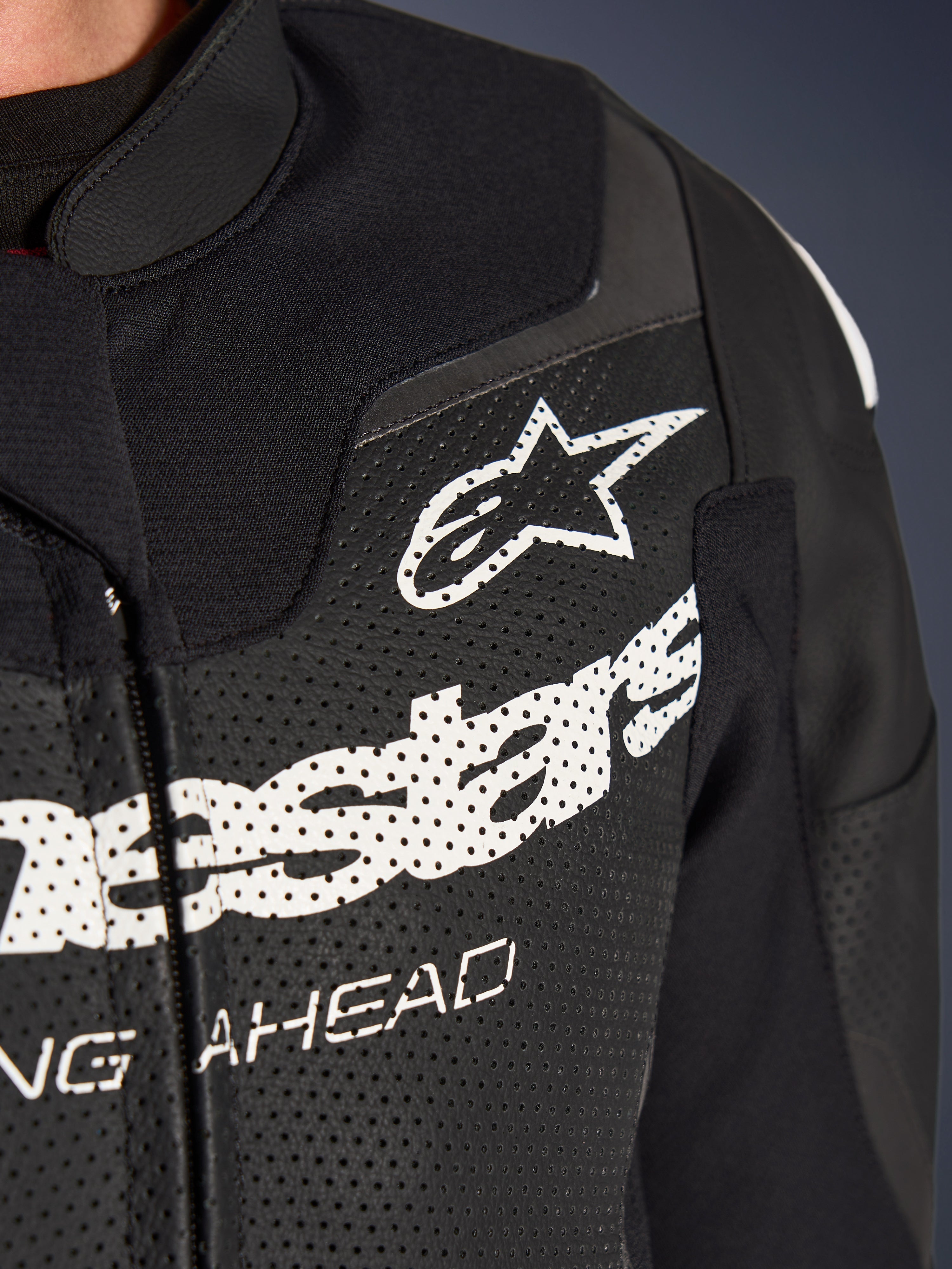 Combinaison en cuir 2 pièces Alpinestars GP Force V2, combinaison de moto de course/sport, noire, gros plan de la poitrine en cuir perforé avec logo et lettrage Alpinestars blancs, soulignant les perforations de ventilation et la construction texturée.