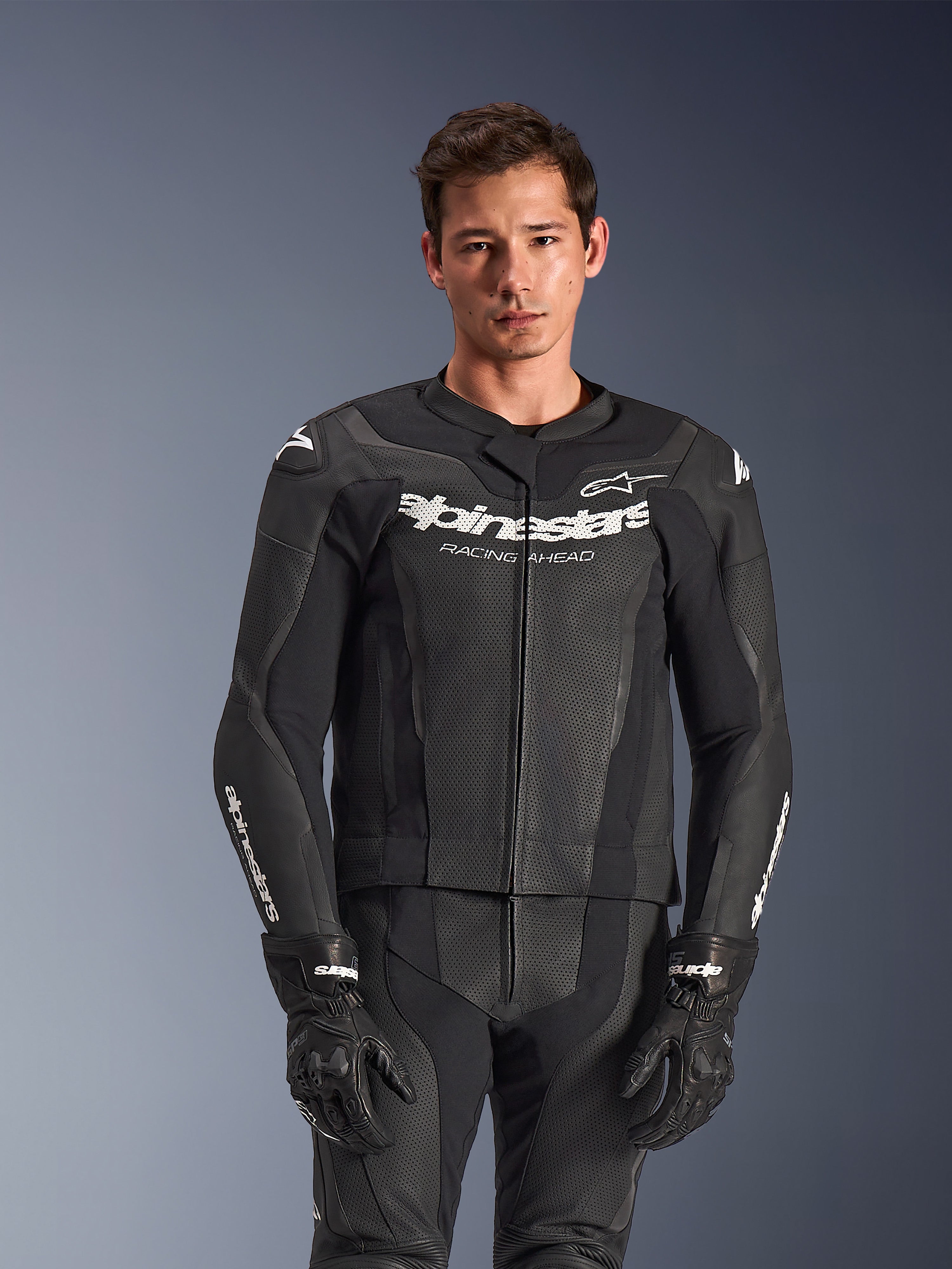 Combinaison 2 pièces en cuir Alpinestars GP Force V2, noire, portée par un mannequin, avec panneaux en cuir perforé et logo blanc 'Alpinestars Racing Ahead' sur la poitrine, gants noirs, équipement de conduite sportive professionnel