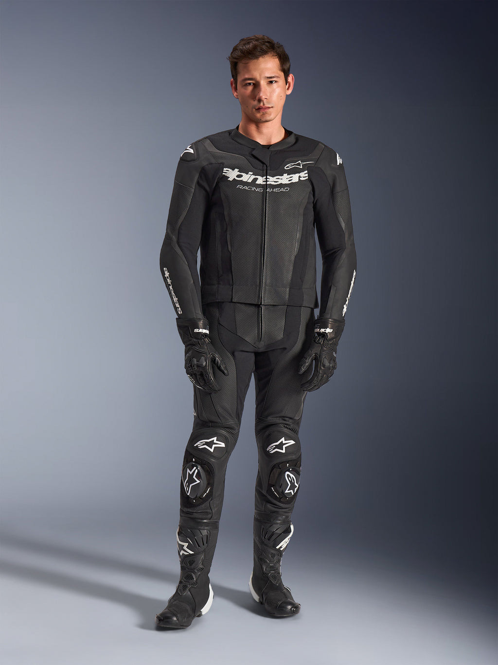 Alpinestars GP Force V2, combinaison de moto en cuir deux pièces, noire avec logos blancs, équipement de course intégral en cuir de qualité avec sliders de genoux renforcés et panneaux perforés, portée avec des gants et des bottes noirs, vue studio en pied