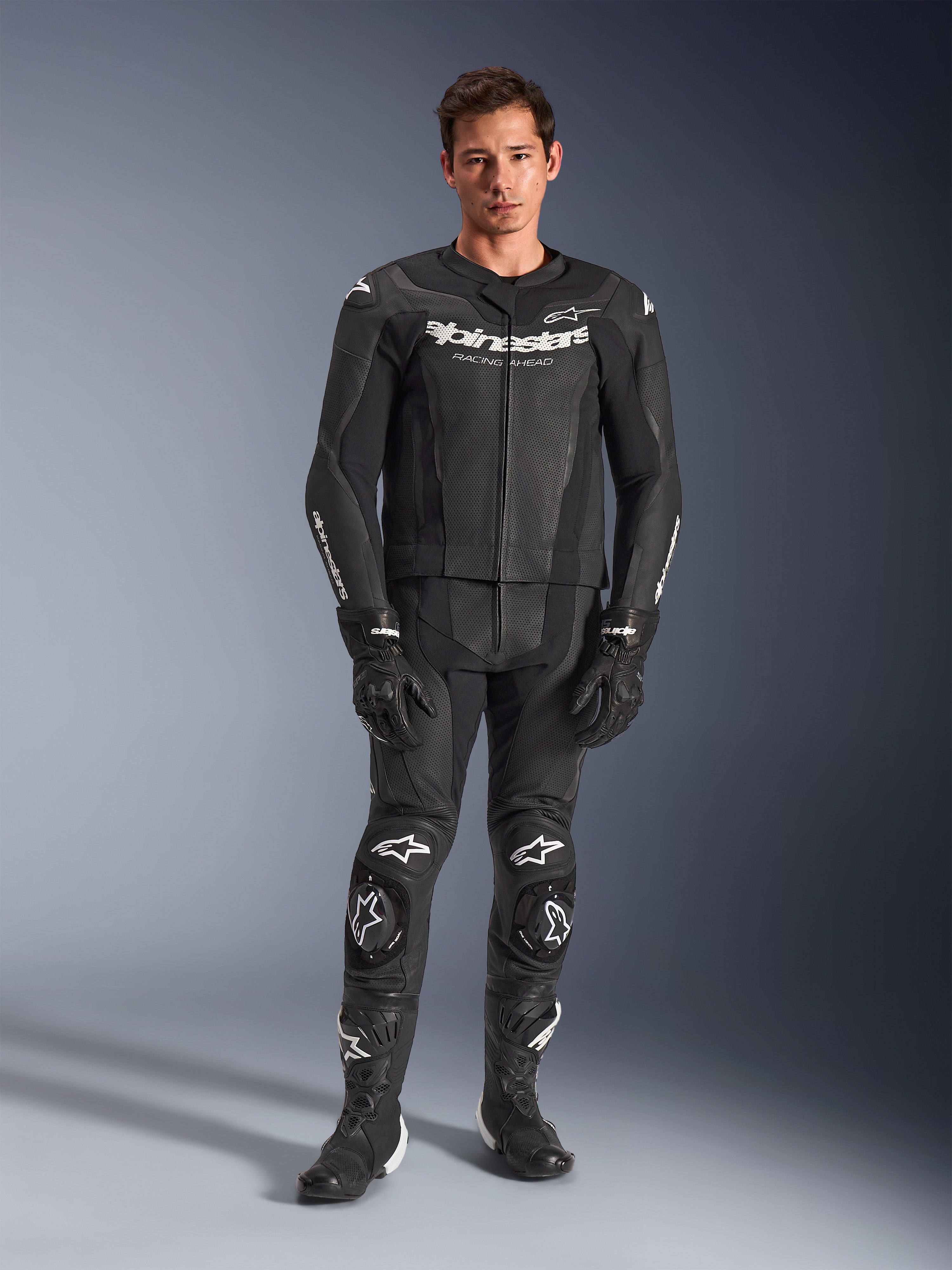 Alpinestars GP Force V2, combinaison de moto en cuir deux pièces, noire avec logos blancs, équipement de course intégral en cuir de qualité avec sliders de genoux renforcés et panneaux perforés, portée avec des gants et des bottes noirs, vue studio en pied