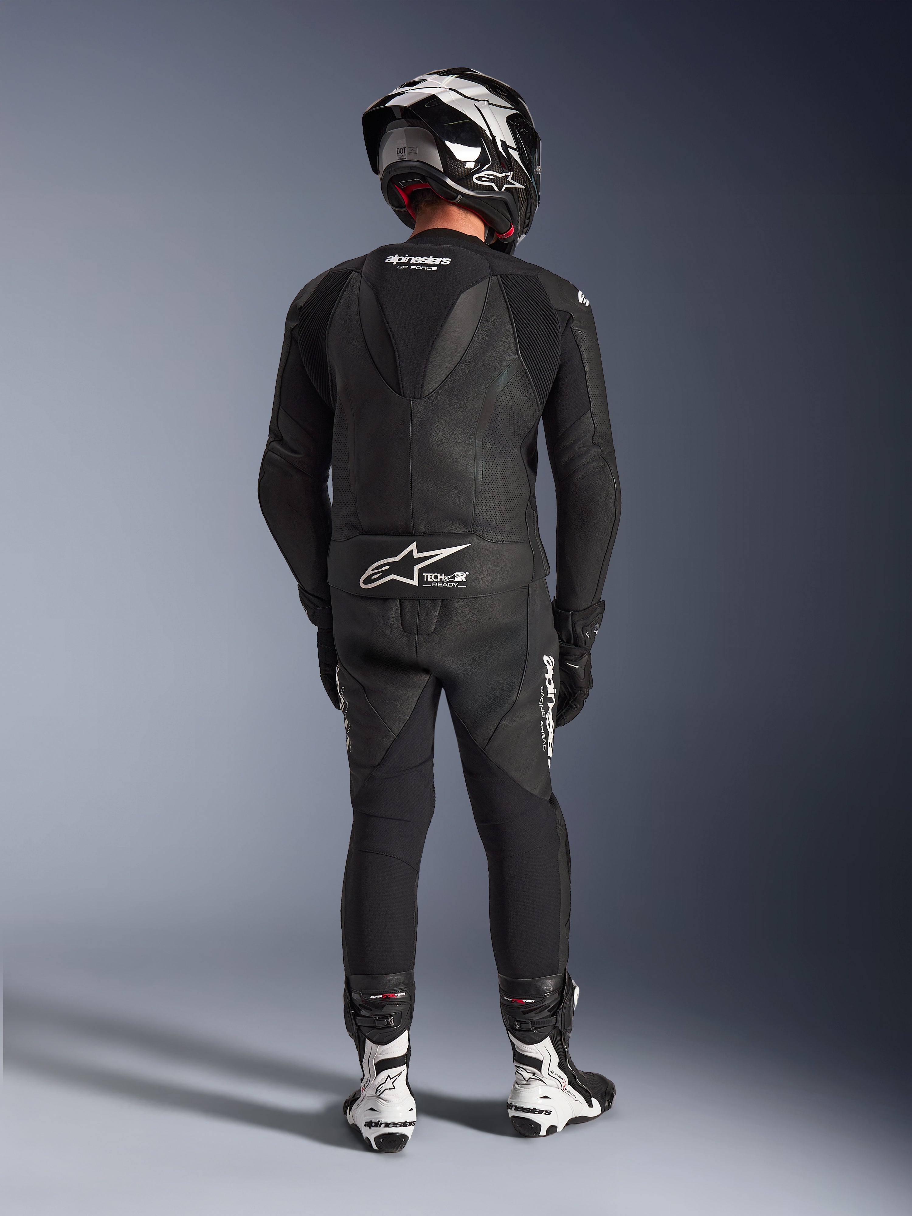 Combinaison en cuir 2 pièces Alpinestars GP Force V2 en noir, vue arrière complète montrant la bosse aérodynamique et le marquage Tech-Air Ready, associée à un casque noir et blanc et des bottes de course blanches pour une performance professionnelle sur circuit