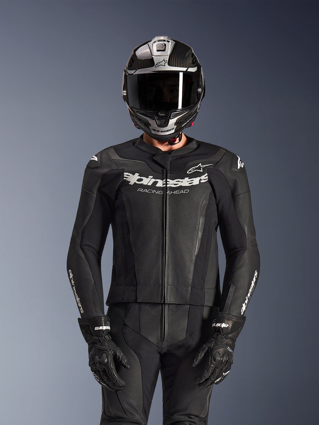 Pilote portant la combinaison en cuir 2 pièces Alpinestars GP Force V2 en noir, construction en cuir perforé avec logo blanc « Racing Ahead » sur la poitrine, casque intégral argenté et en fibre de carbone, gants noirs, équipement professionnel pour la conduite sportive et sur circuit