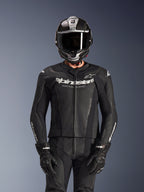 Pilote portant la combinaison en cuir 2 pièces Alpinestars GP Force V2 en noir, construction en cuir perforé avec logo blanc « Racing Ahead » sur la poitrine, casque intégral argenté et en fibre de carbone, gants noirs, équipement professionnel pour la conduite sportive et sur circuit