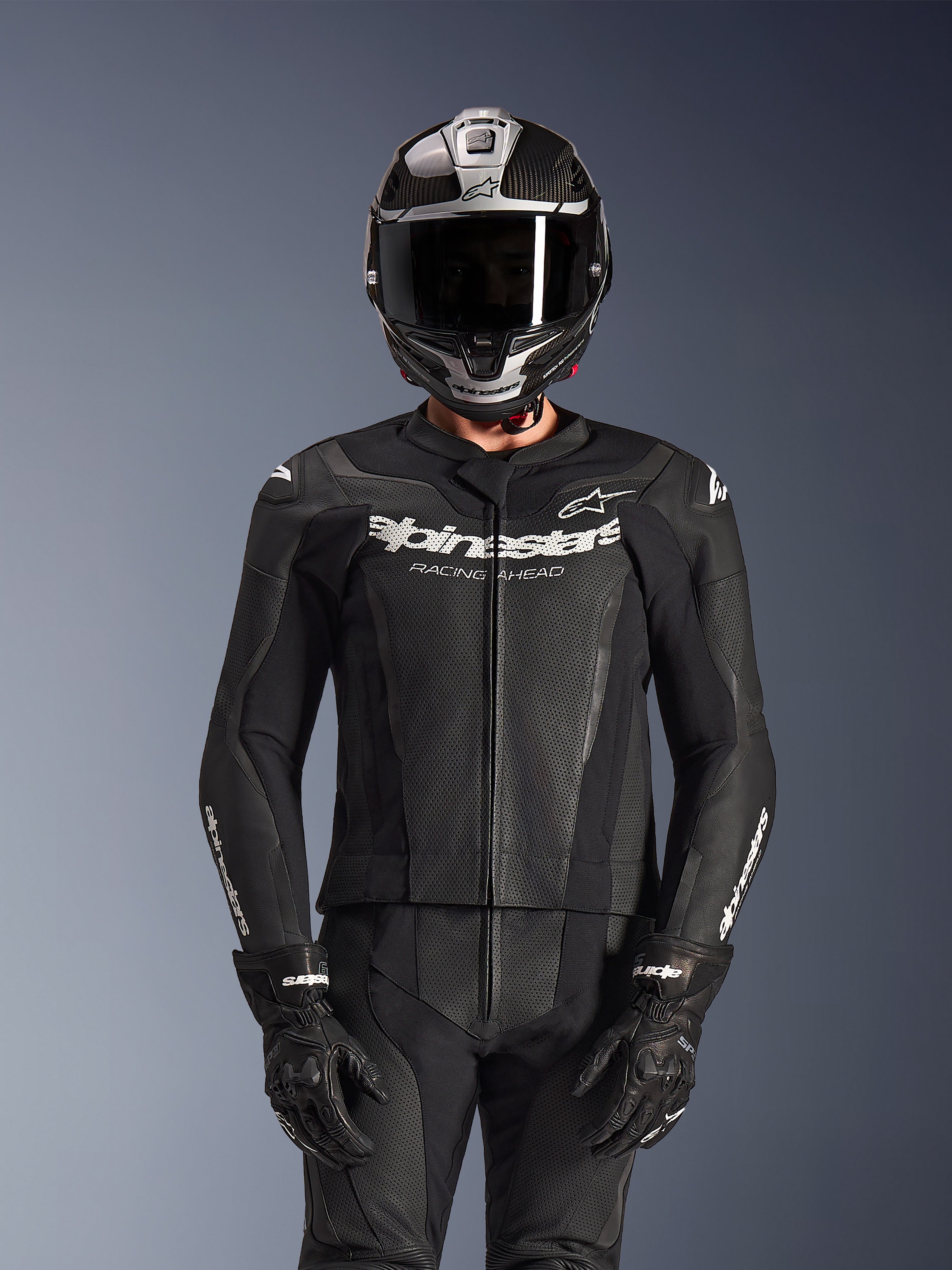 Pilote portant la combinaison en cuir 2 pièces Alpinestars GP Force V2 en noir, construction en cuir perforé avec logo blanc « Racing Ahead » sur la poitrine, casque intégral argenté et en fibre de carbone, gants noirs, équipement professionnel pour la conduite sportive et sur circuit