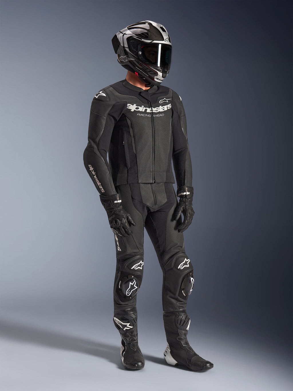 Combinaison en cuir 2 pièces Alpinestars GP Force V2, combinaison de course de moto noire, portée avec un casque intégral, des gants et des bottes, dotée d'une construction en cuir perforé, d'une bosse aérodynamique et de logos Alpinestars blancs sur la poitrine, les bras et les genoux.