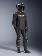 Combinaison en cuir 2 pièces Alpinestars GP Force V2, combinaison de course de moto noire, portée avec un casque intégral, des gants et des bottes, dotée d'une construction en cuir perforé, d'une bosse aérodynamique et de logos Alpinestars blancs sur la poitrine, les bras et les genoux.