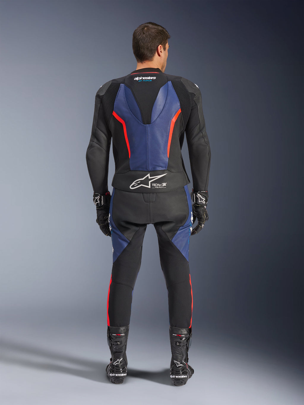 Combinaison en cuir Alpinestars GP Force V2 2 pièces, vue arrière, noir, bleu et rouge fluo, avec des panneaux en cuir bleu sur le dos et les jambes et des accents rouge fluo, marquage Tech-Air Ready en bas du dos, portée avec des gants et bottes de course noirs.