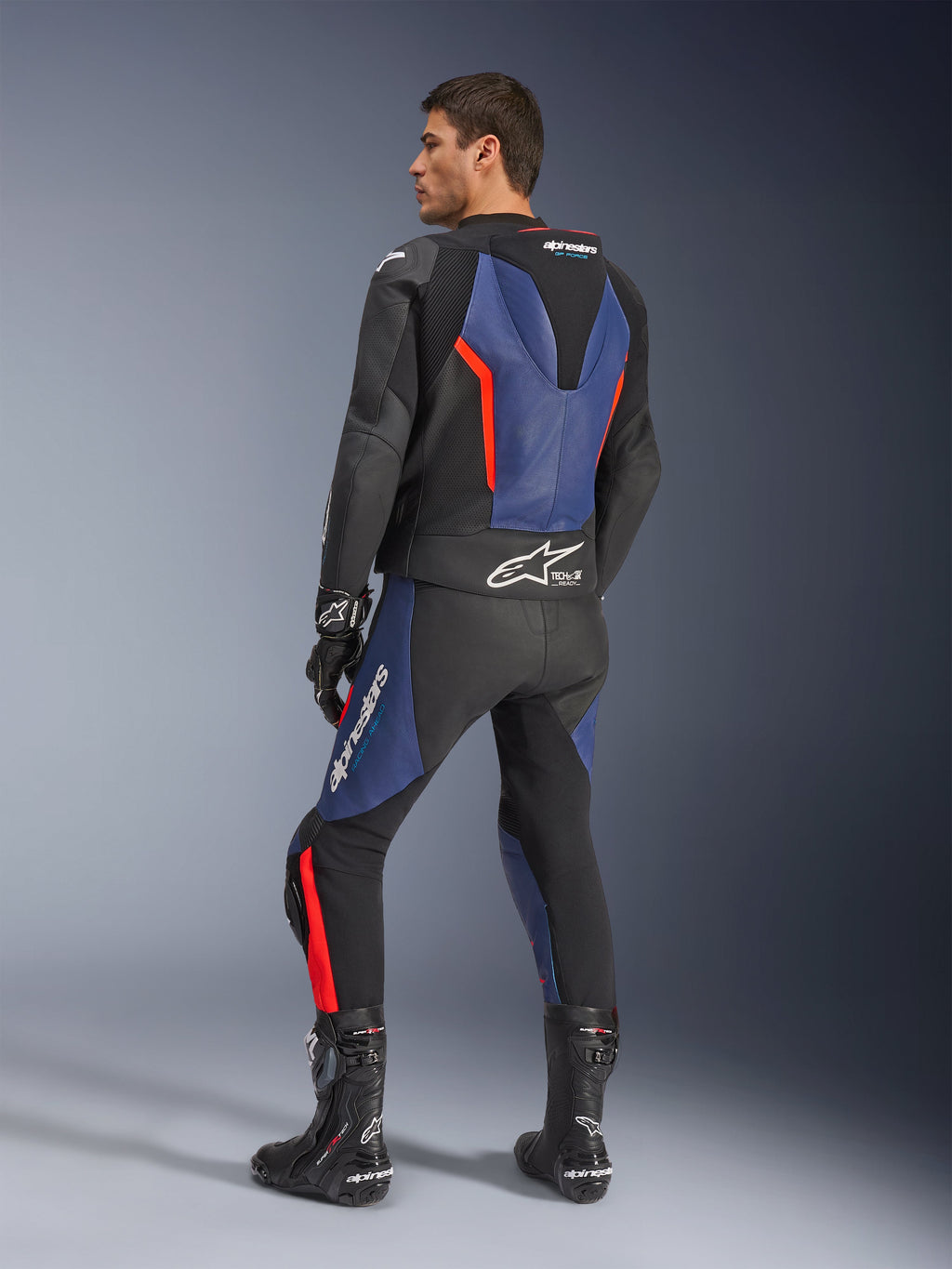 Combinaison en cuir 2 pièces GP Force V2, noir, bleu et rouge fluo, vue de profil arrière sur un mannequin, avec bosse aérodynamique, cuir perforé et logo Tech-Air Ready, portée avec des bottes et gants de course Alpinestars noirs