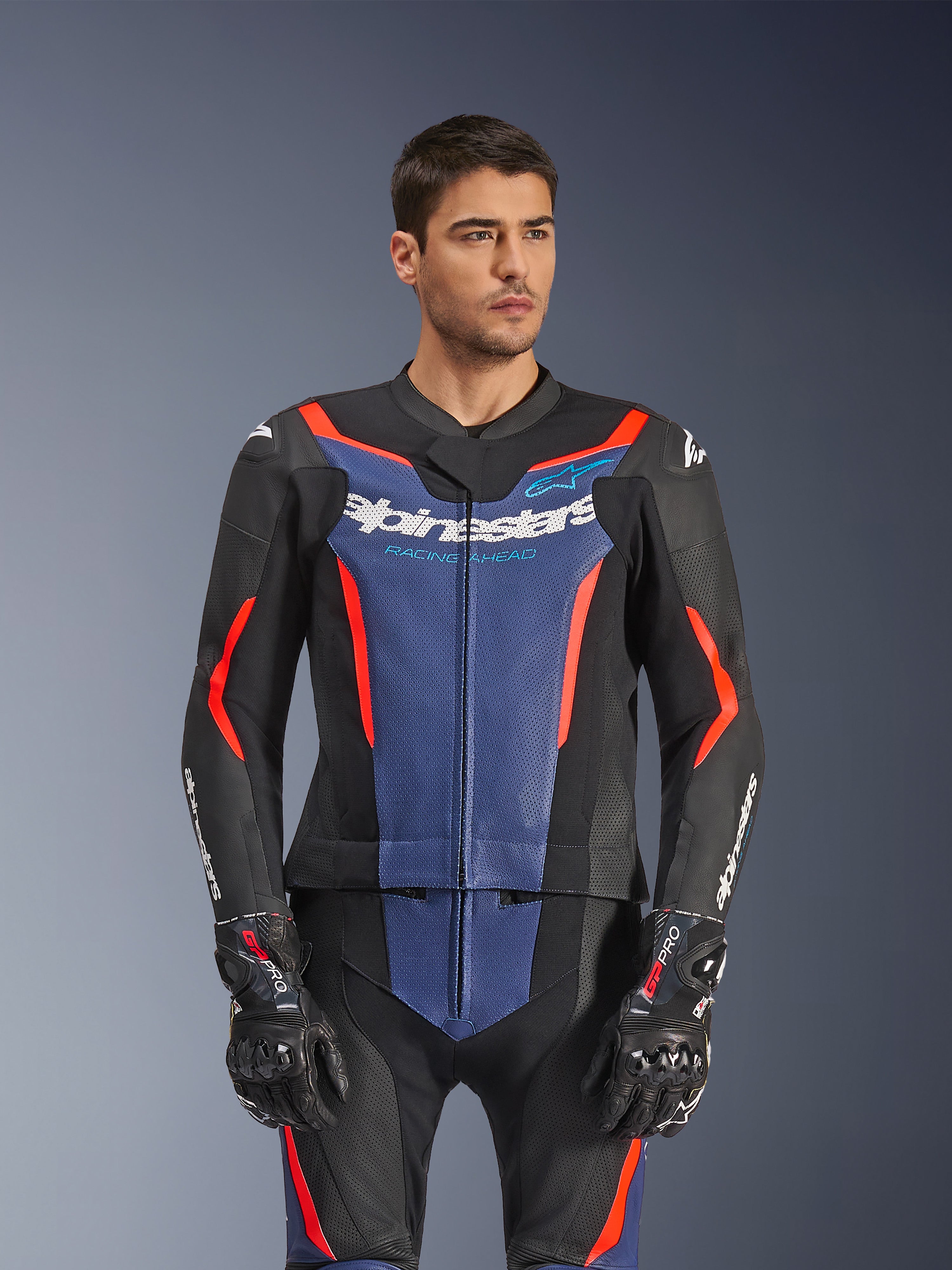 Modèle masculin portant la combinaison en cuir Alpinestars GP Force V2 2 pièces, noir bleu rouge fluo, avec un empiècement bleu sur la poitrine et des accents rouges, portée avec des gants GP Pro, équipement professionnel de course moto en cuir avec texture perforée.