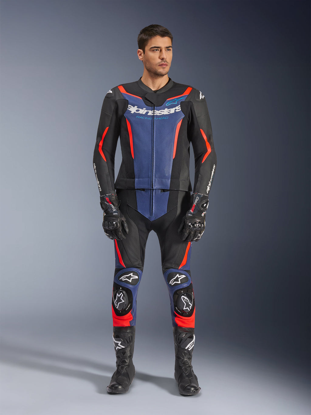 Alpinestars GP Force V2 Leather Suit 2-Piece en noir, bleu et rouge fluo, dotée de panneaux bleus sur la poitrine et le bas des jambes avec des accents rouge fluo éclatants, portée par un mannequin homme avec des bottes et des gants de course noirs, équipement de protection professionnel pour le sport moto