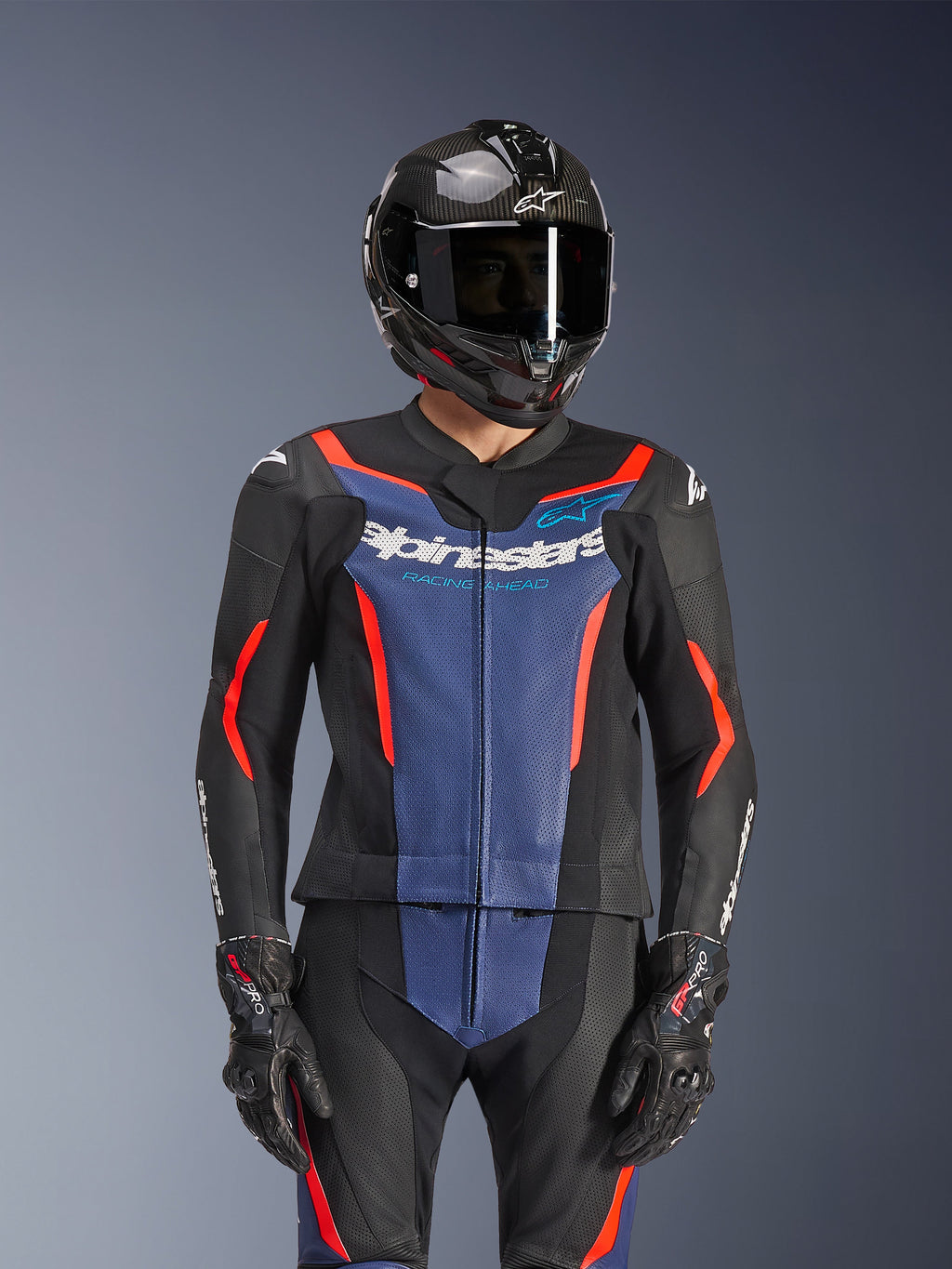 Combinaison en cuir 2 pièces Alpinestars GP Force V2, noir, bleu et rouge fluo, portée avec un casque en fibre de carbone et des gants de course, avec panneaux en cuir perforé et logo Alpinestars blanc sur la poitrine, équipement de course professionnel et de conduite sportive