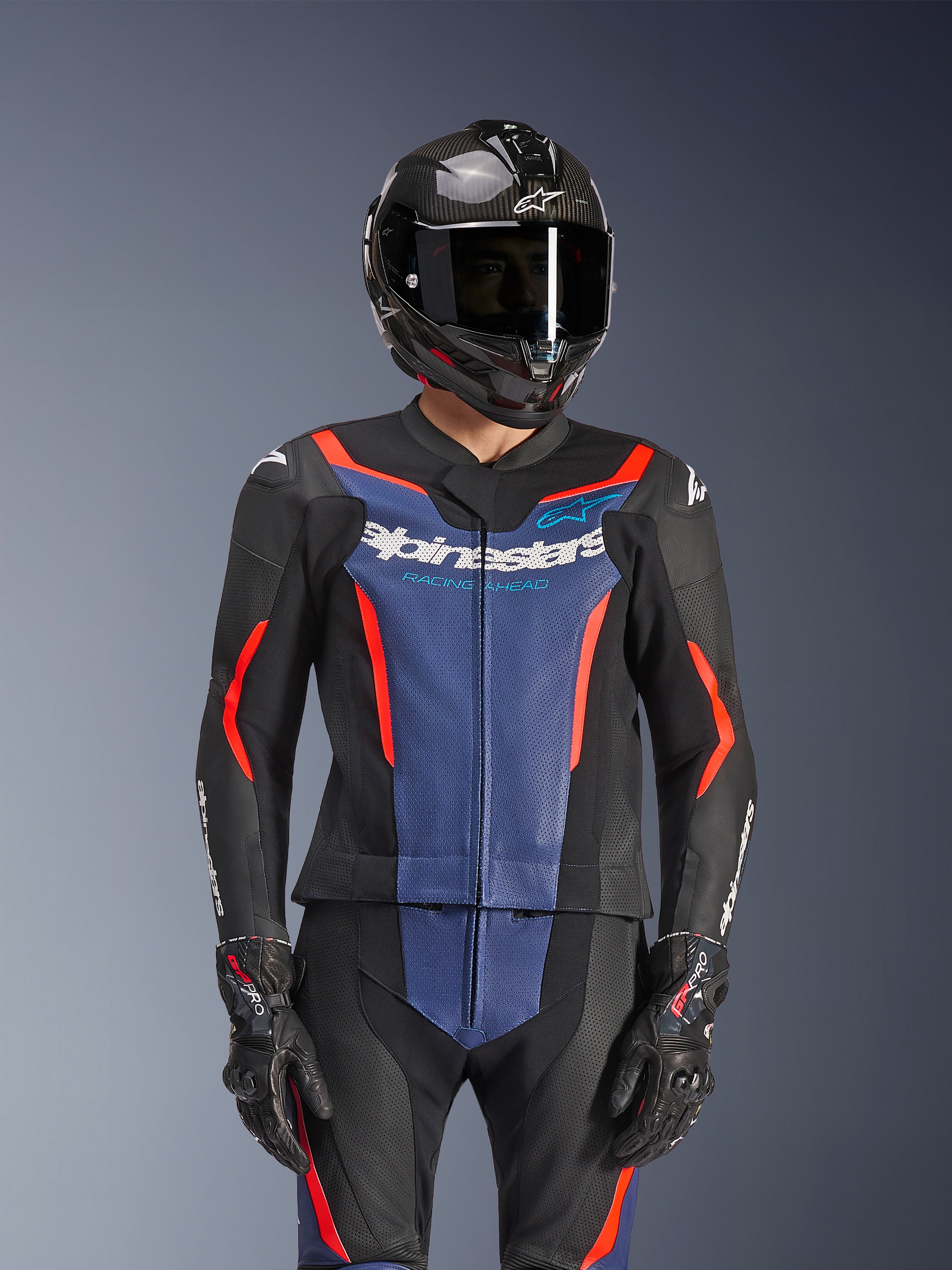 Combinaison en cuir 2 pièces Alpinestars GP Force V2, noir, bleu et rouge fluo, portée avec un casque en fibre de carbone et des gants de course, avec panneaux en cuir perforé et logo Alpinestars blanc sur la poitrine, équipement de course professionnel et de conduite sportive