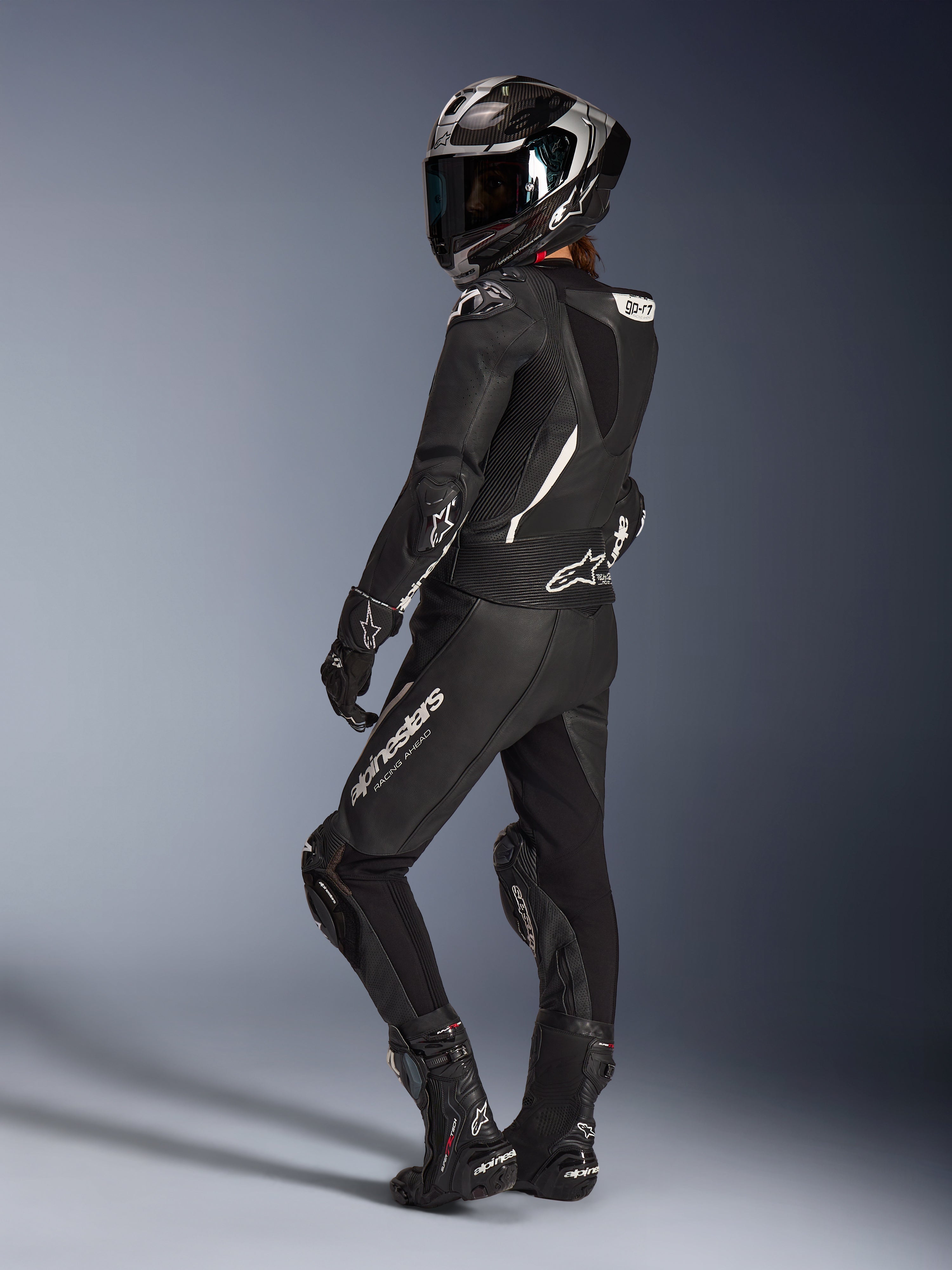 Pilote femme portant la combinaison en cuir 2 pièces Alpinestars Stella GP-R7 en noir, casque de course intégral avec finition carbone, gants et bottes assortis, vue arrière mettant en valeur la bosse aérodynamique et les logos Alpinestars blancs, équipement professionnel pour la piste et la conduite sportive