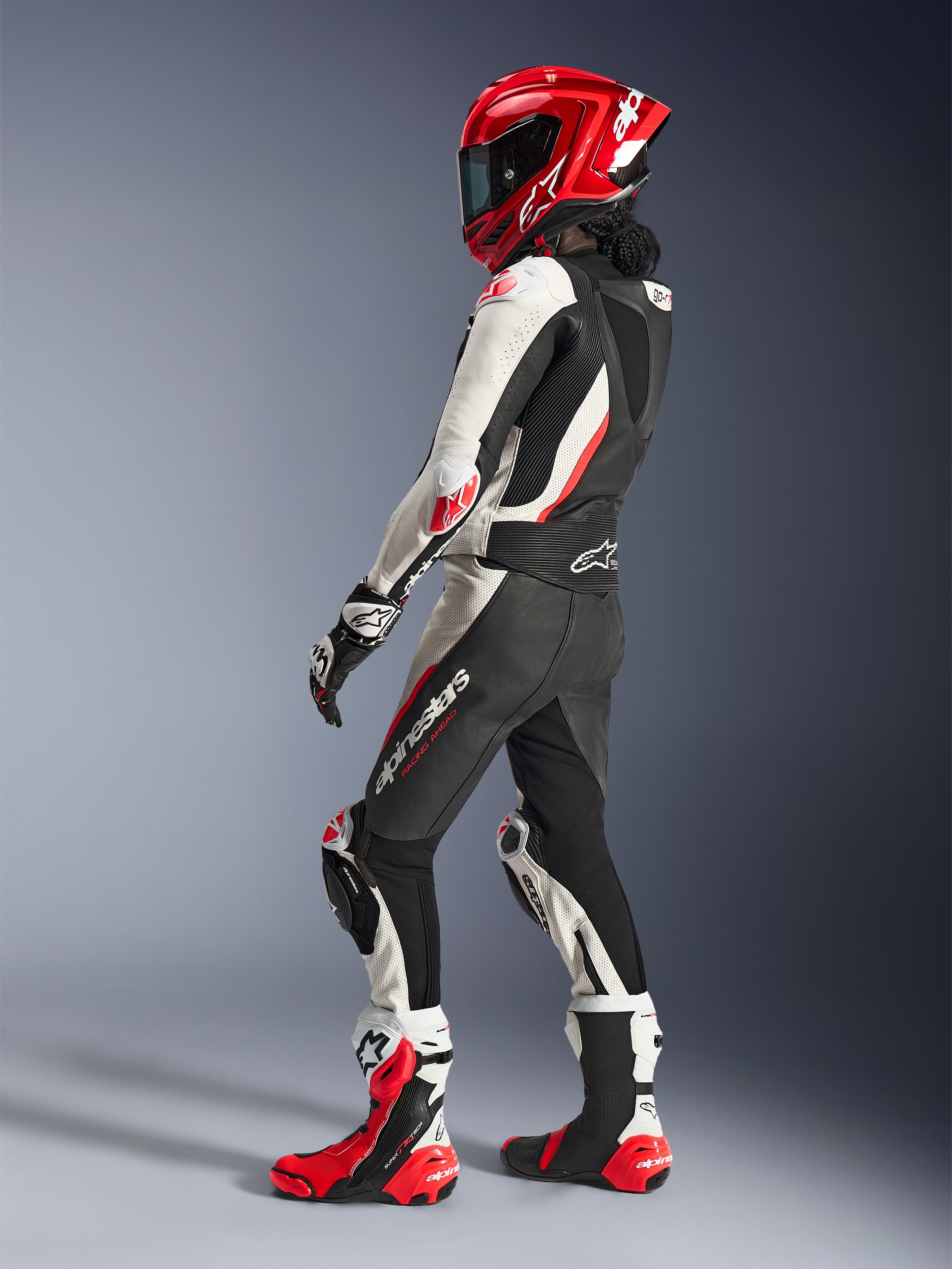 Pilote femme portant la combinaison en cuir 2 pièces Alpinestars Stella GP-R7, noir, blanc et rouge fluo, vue de trois-quarts arrière montrant la bosse aérodynamique, casque intégral rouge assorti, gants de course noirs et blancs et bottes de course techniques rouges et blanches, équipement de moto professionnel