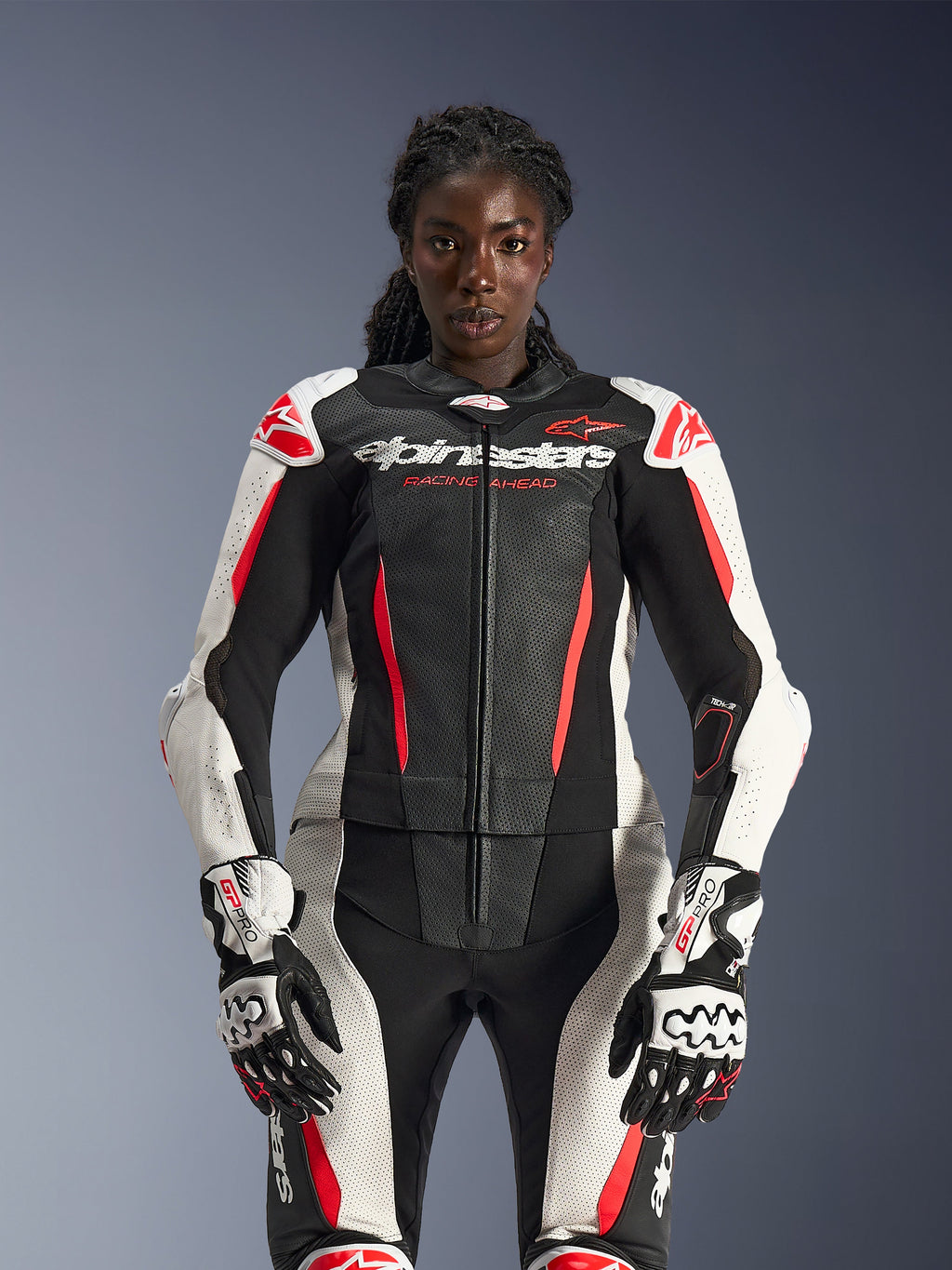 Modèle féminin portant la combinaison en cuir 2 pièces Alpinestars Stella GP-R7 en noir, blanc et rouge fluo, avec des panneaux en cuir perforé et des gants GP Pro assortis, anatomiquement optimisée pour les femmes.