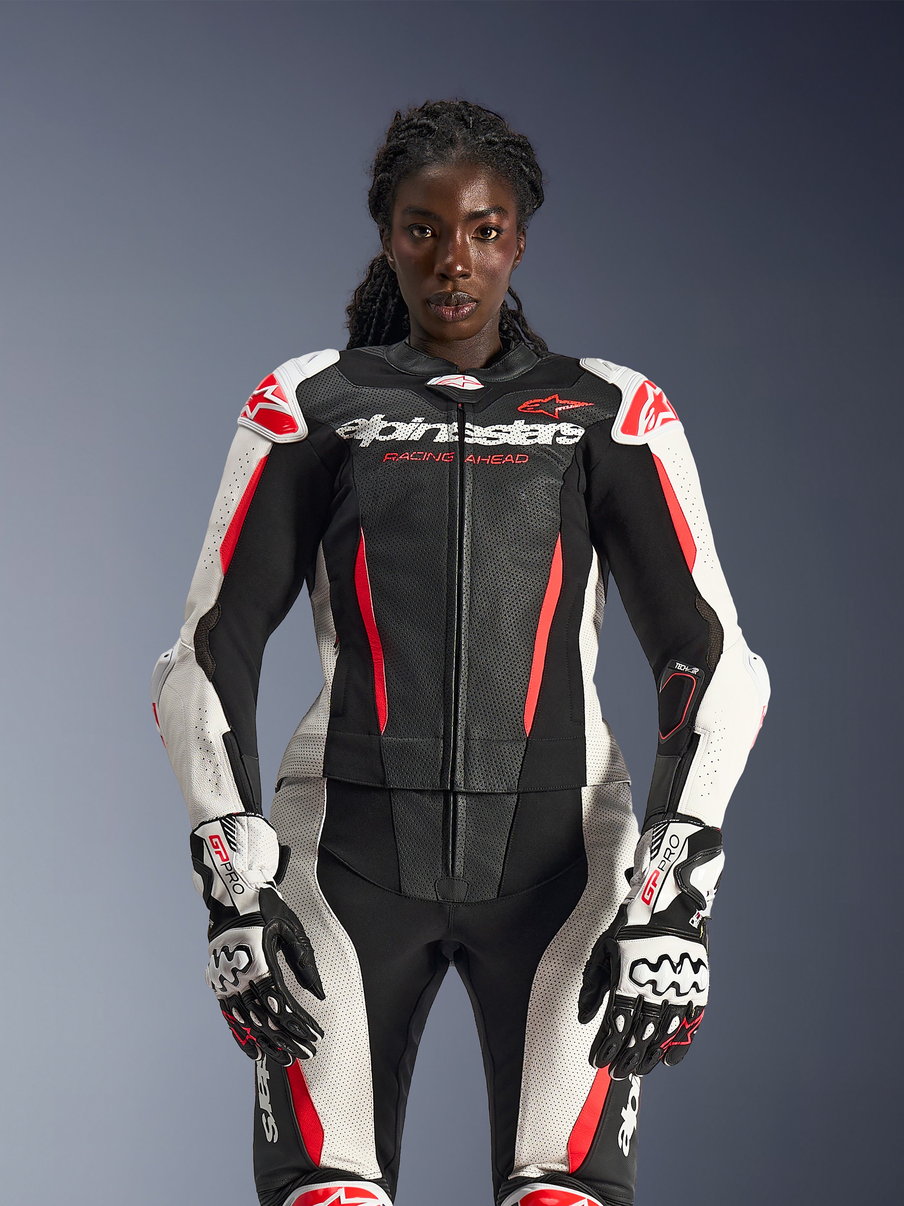 Modèle féminin portant la combinaison en cuir 2 pièces Alpinestars Stella GP-R7 en noir, blanc et rouge fluo, avec des panneaux en cuir perforé et des gants GP Pro assortis, anatomiquement optimisée pour les femmes.