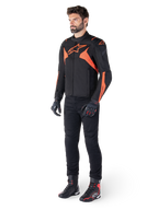 Veste imperméable Alpinestars T-Jaws V4, noir et rouge fluo, portée par un mannequin homme avec pantalon, gants et bottes noirs, avec des accents orange vif et le logo Alpinestars, design sportif pour la conduite de moto