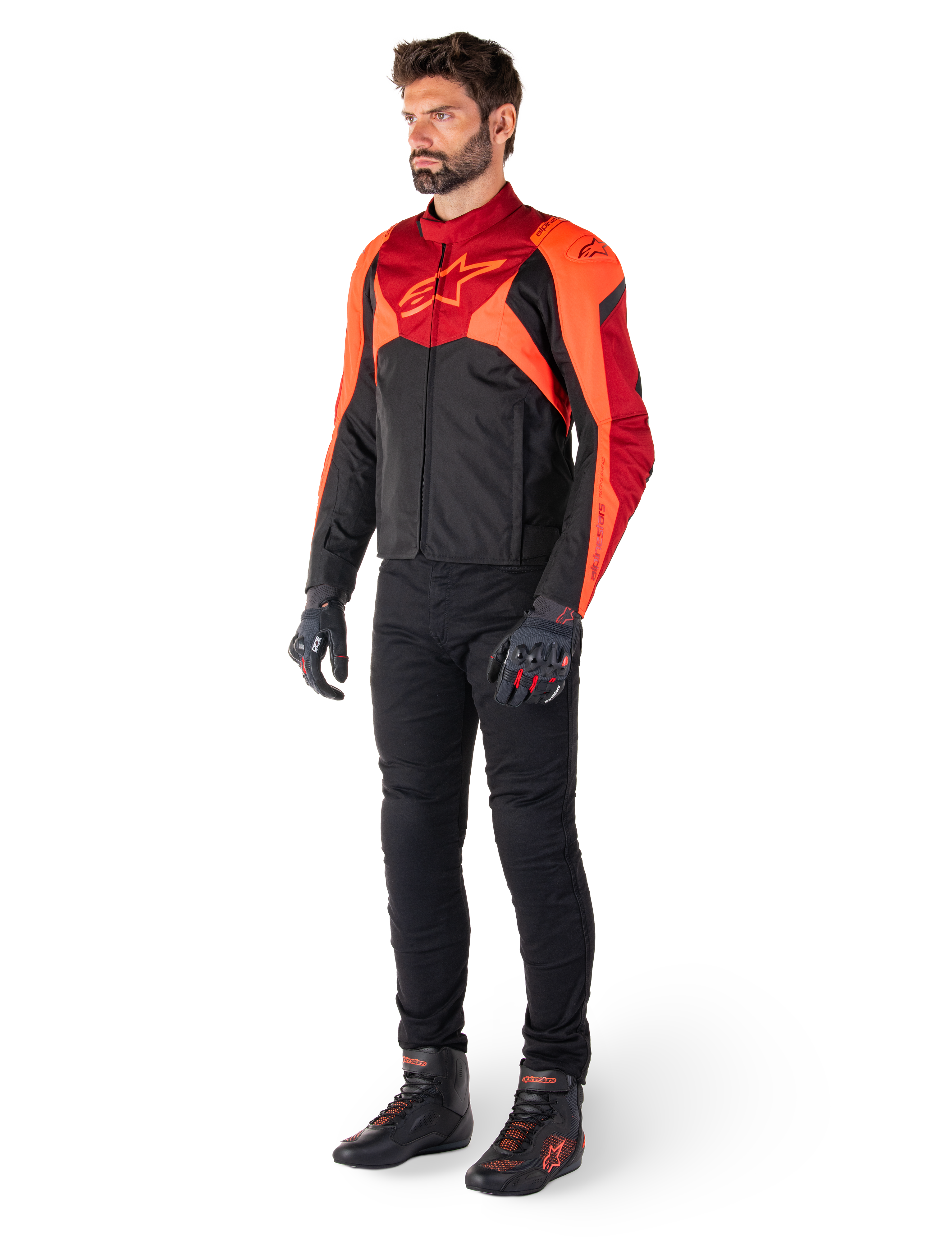 Male rider wearing the Alpinestars T-Jaws V4 Waterproof Jacket in Black Red Fluo Dark Red, paired with black motorcycle pants, gloves, and riding boots, featuring a sport-fit textile design with bold chest branding and an aggressive silhouette.