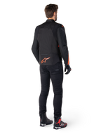 Veste imperméable Alpinestars T-Jaws V4, noir et rouge fluo, vue de dos portée par un motard, présentant un logo rouge fluo éclatant dans le dos, associée à un pantalon de moto noir et des bottes techniques, équipement de moto sportif.