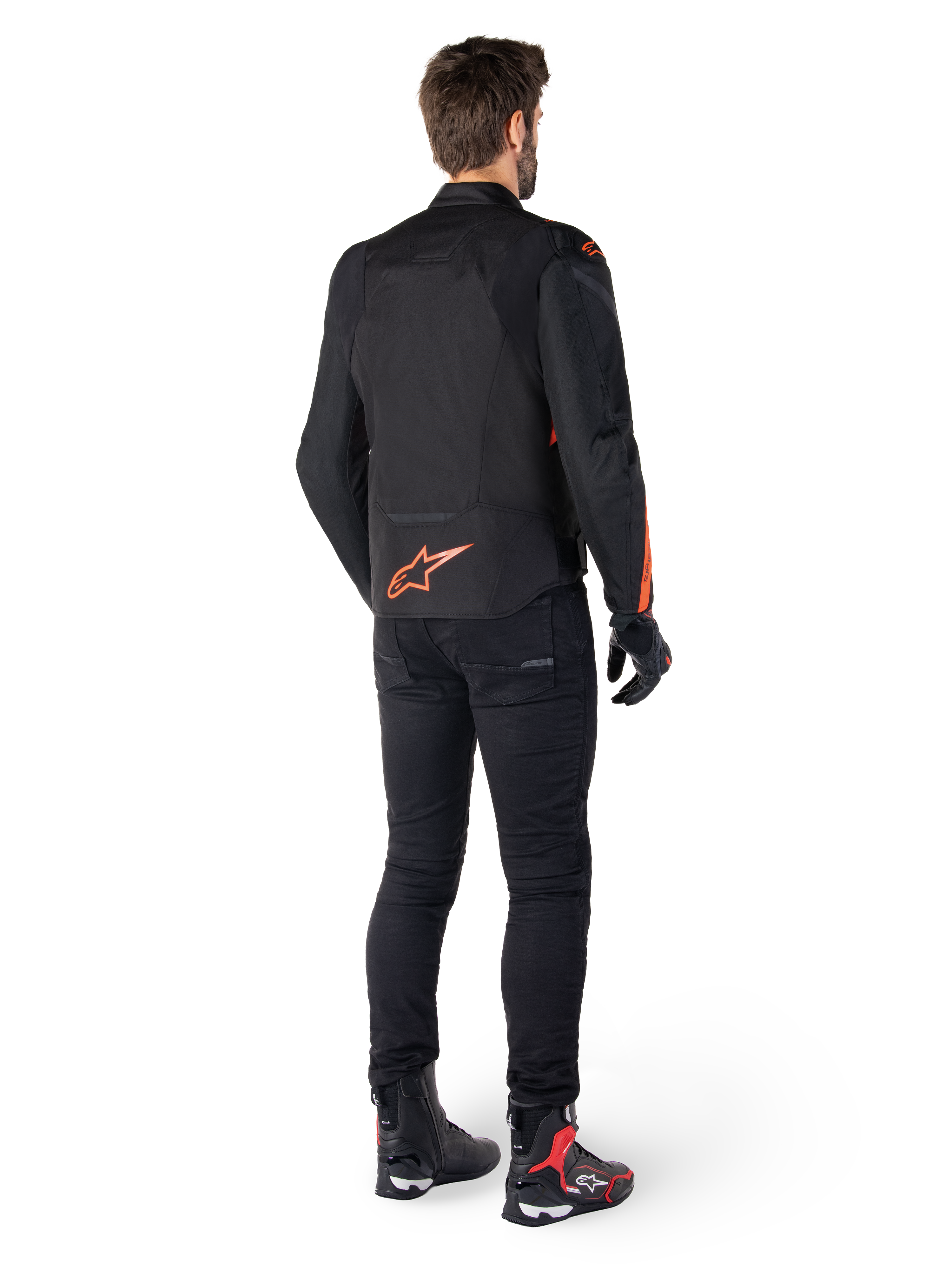 Veste imperméable Alpinestars T-Jaws V4, noir et rouge fluo, vue de dos portée par un motard, présentant un logo rouge fluo éclatant dans le dos, associée à un pantalon de moto noir et des bottes techniques, équipement de moto sportif.