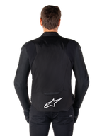 Alpinestars T-Jaws V4 Waterproof Jacket, veste de moto noire vue de dos, arborant un logo Alpinestars blanc sur le bas du dos, dos rallongé pour une meilleure protection, portée par un mannequin avec pantalon et gants noirs, design textile sport épuré