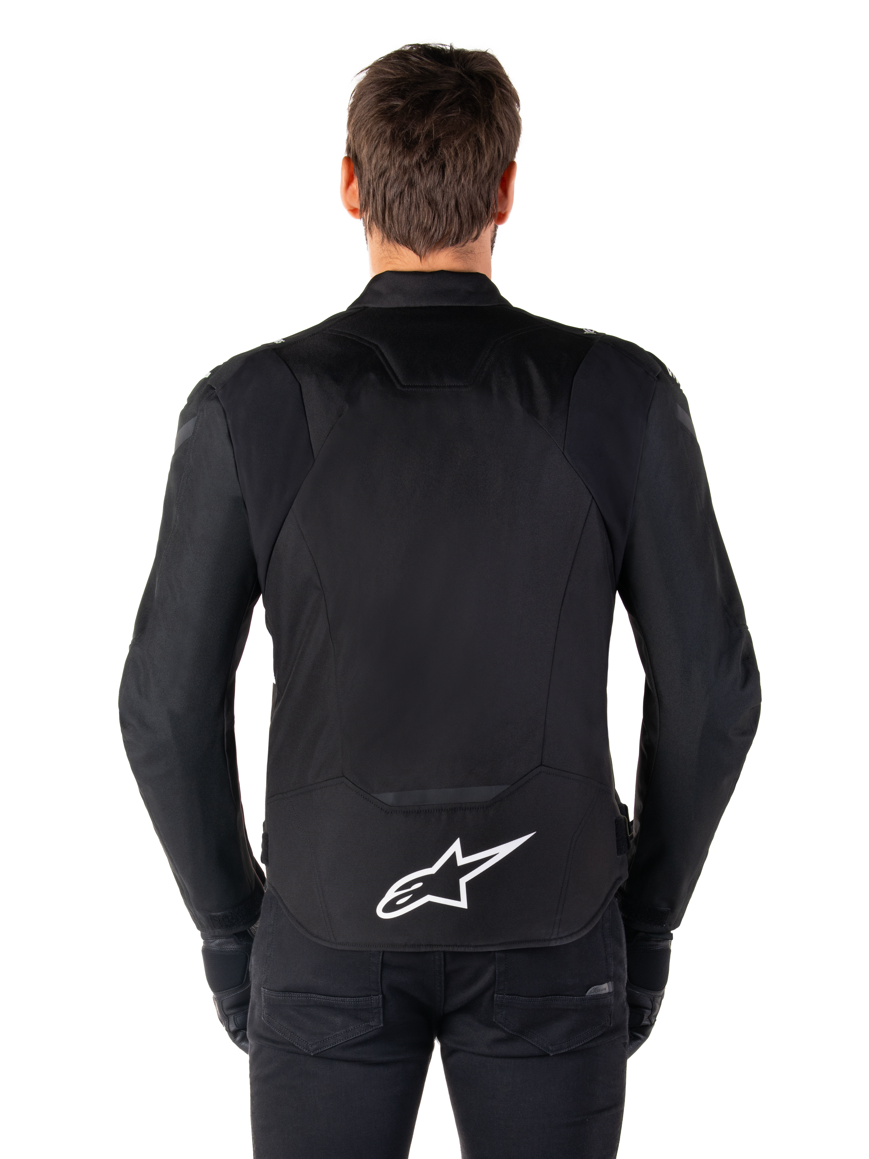 Alpinestars T-Jaws V4 Waterproof Jacket, veste de moto noire vue de dos, arborant un logo Alpinestars blanc sur le bas du dos, dos rallongé pour une meilleure protection, portée par un mannequin avec pantalon et gants noirs, design textile sport épuré