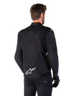 Veste étanche Alpinestars T-Jaws V4, noir et blanc, vue arrière, veste de moto sport en textile avec logo blanc sur le bas du dos, portée avec des gants et un pantalon noirs, présentant une silhouette à coupe sport révisée