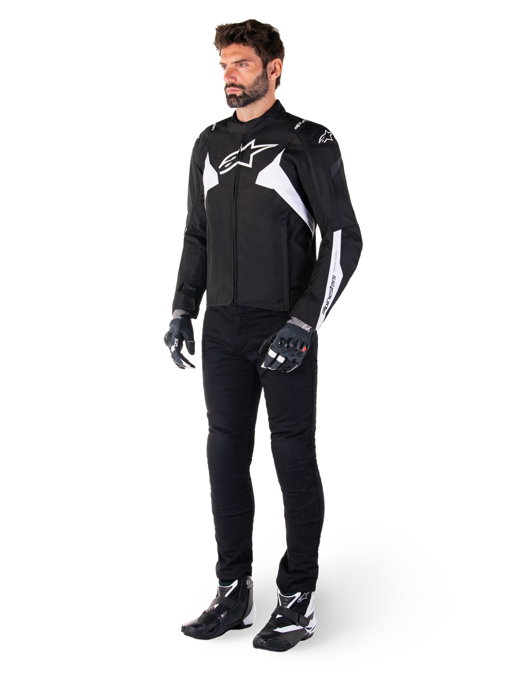 Modèle masculin portant la veste imperméable Alpinestars T-Jaws V4 en noir et blanc, veste sport en textile arborant un logo d'étoile blanche et des accents sur les manches, associée à un pantalon de moto noir, des gants et des bottes pour un look de route complet.