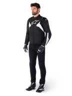 Modèle masculin portant la veste imperméable Alpinestars T-Jaws V4 en noir et blanc, veste sport en textile arborant un logo d'étoile blanche et des accents sur les manches, associée à un pantalon de moto noir, des gants et des bottes pour un look de route complet.
