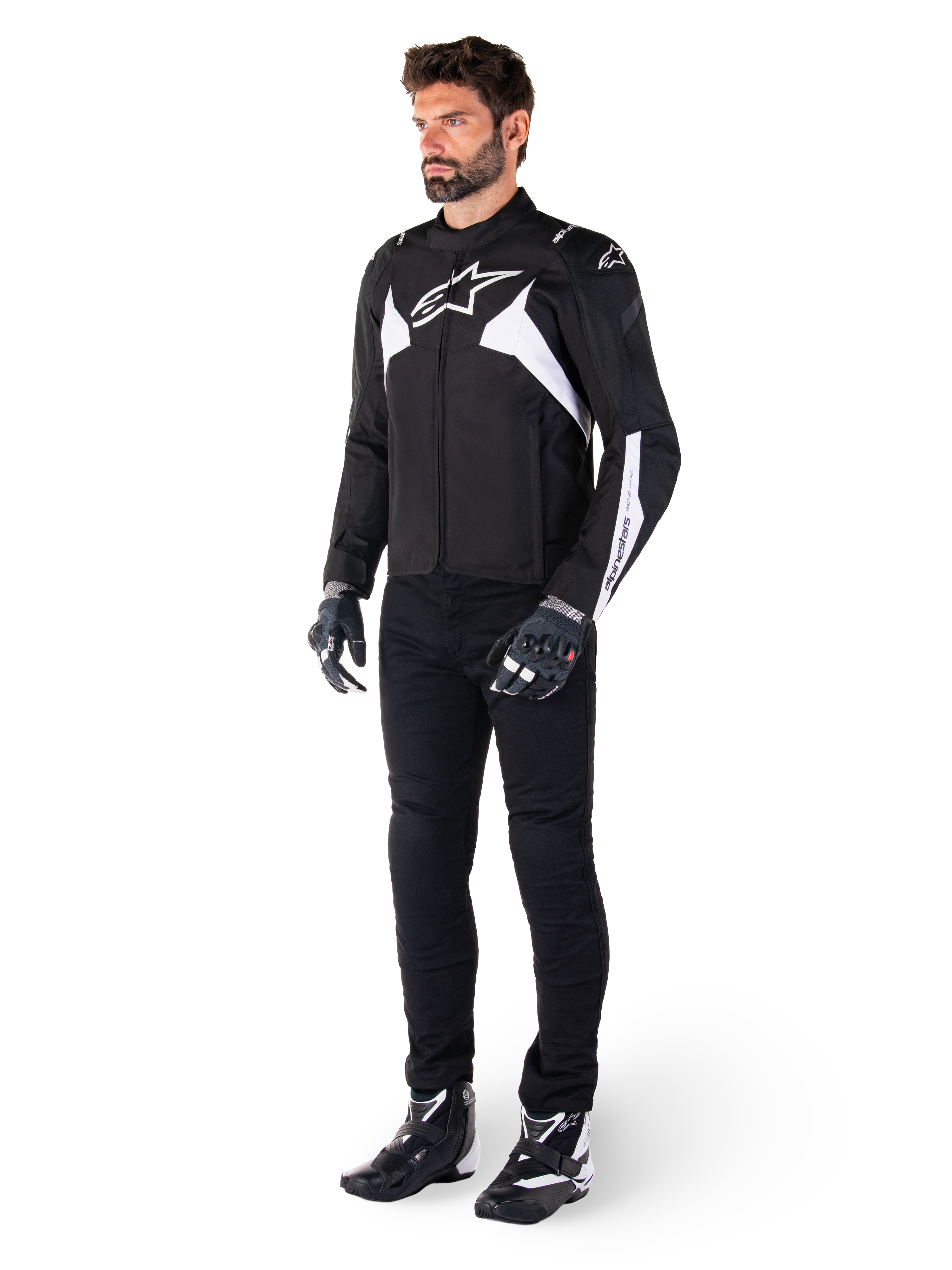 Modèle masculin portant la veste imperméable Alpinestars T-Jaws V4 en noir et blanc, veste sport en textile arborant un logo d'étoile blanche et des accents sur les manches, associée à un pantalon de moto noir, des gants et des bottes pour un look de route complet.