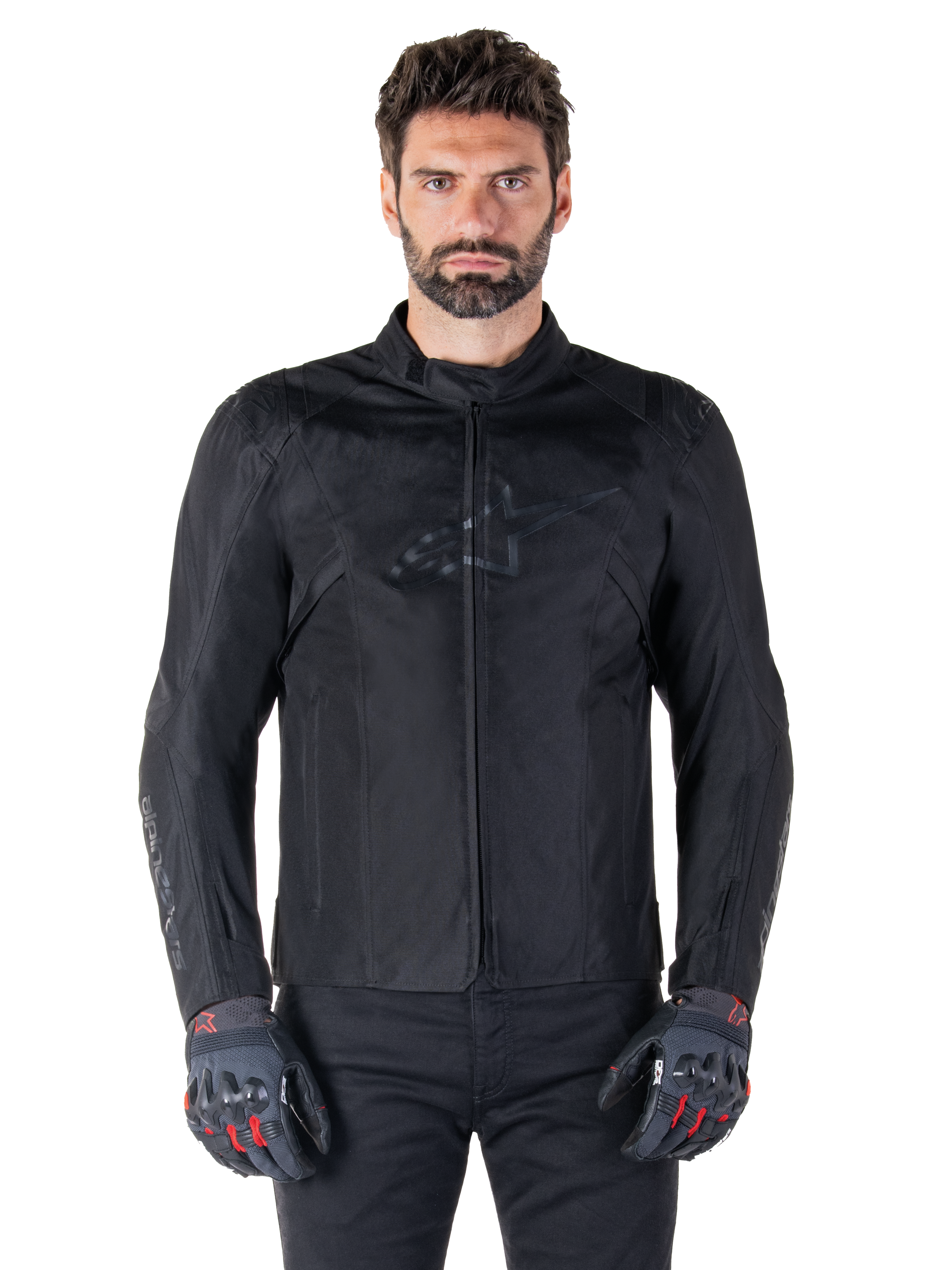 Alpinestars T-SPS V2 Waterproof Jacket, veste de moto textile noire, portée par un motard avec des gants noirs et rouges, dotée d'un col montant, d'une fermeture éclair frontale et d'un logo discret sur la poitrine, protection imperméable pour la conduite sportive et urbaine