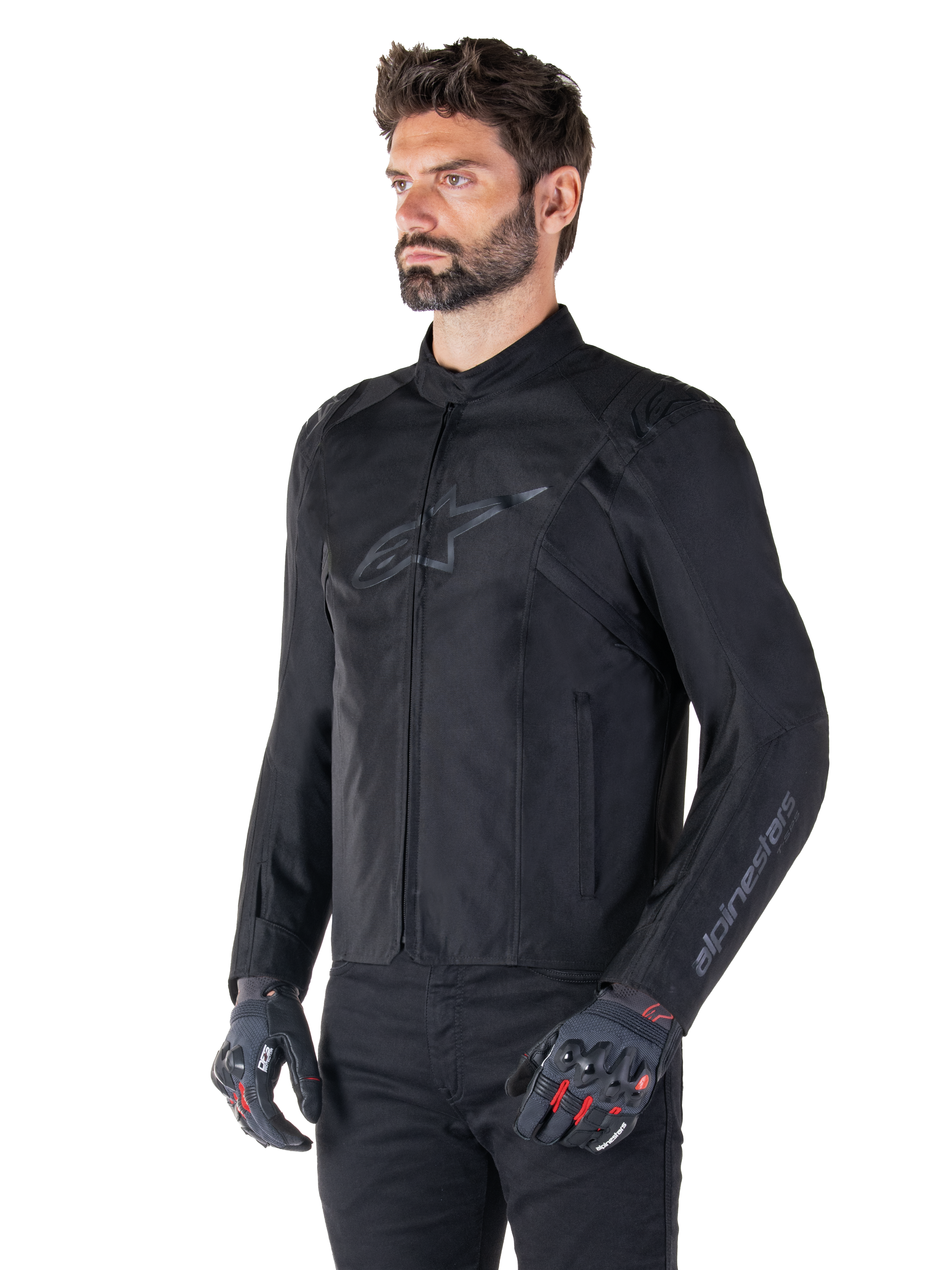 Veste Alpinestars T-SPS V2 Waterproof, noir, portée par un mannequin avec un pantalon noir et des gants de moto gris avec accents rouges, dotée d'un logo discret sur la poitrine et d'un marquage sur la manche, veste textile à coupe sport pour la route.