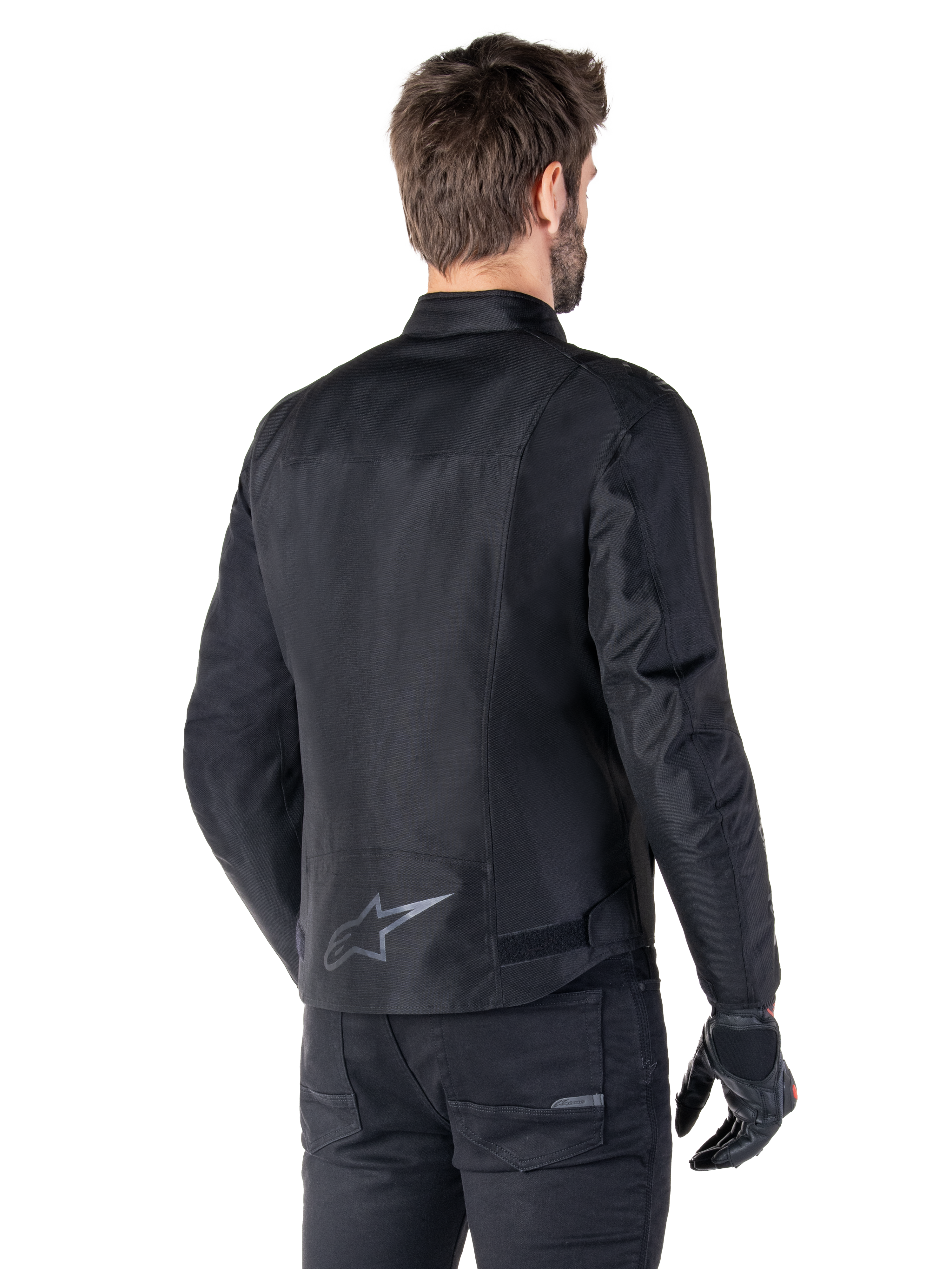 Veste imperméable Alpinestars T-SPS V2, noire, vue arrière sur un mannequin homme, avec un logo étoile réfléchissant dans le bas du dos, un profil arrière allongé et des sangles de réglage à la taille, portée avec un pantalon de moto et des gants noirs