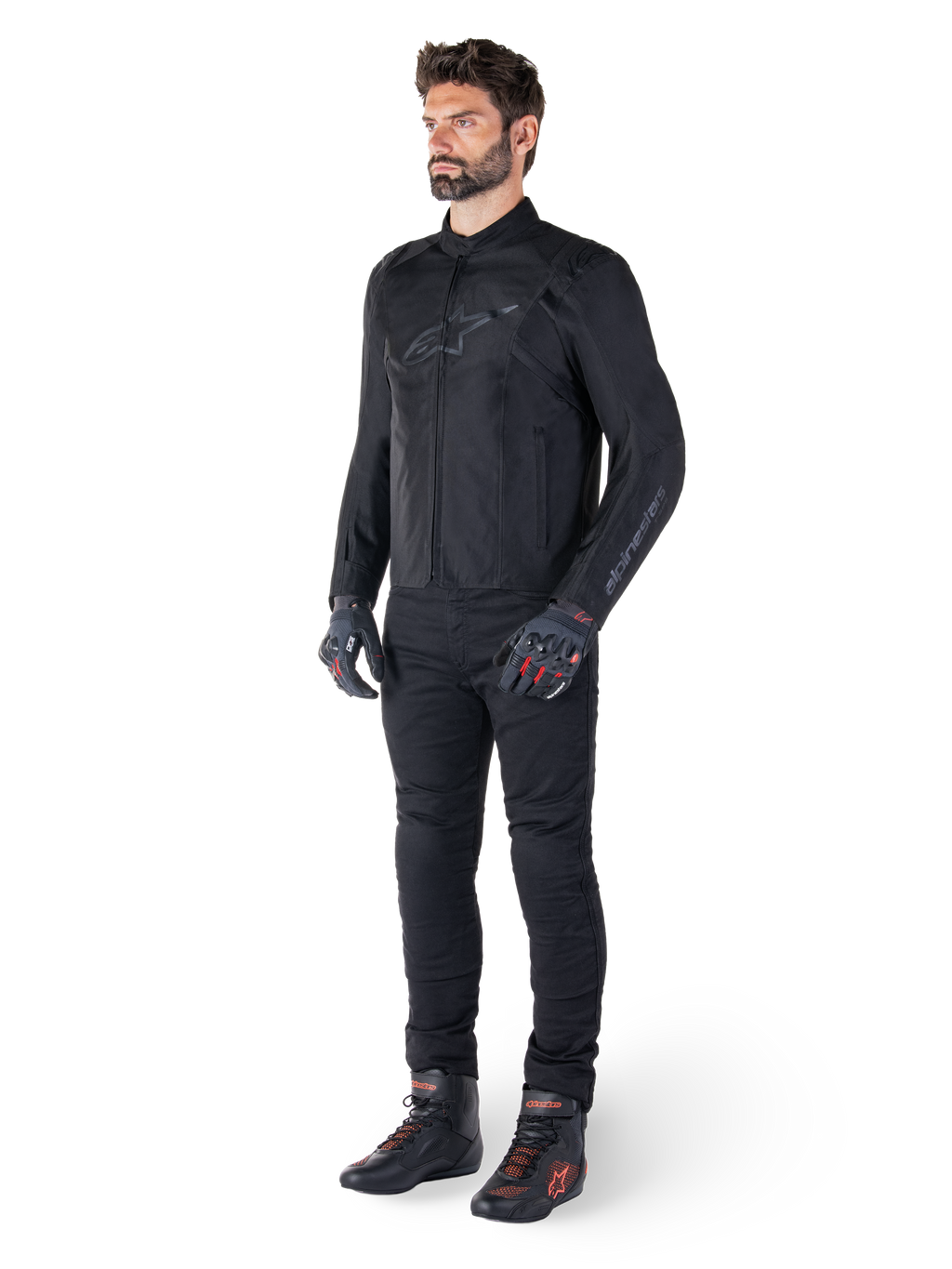 Veste Alpinestars T-SPS V2 Waterproof, noire, portée par un mannequin homme avec un pantalon noir assorti, des gants noirs avec des accents rouges et des chaussures de moto noires, équipement de moto complet pour la conduite urbaine et sportive, fond blanc