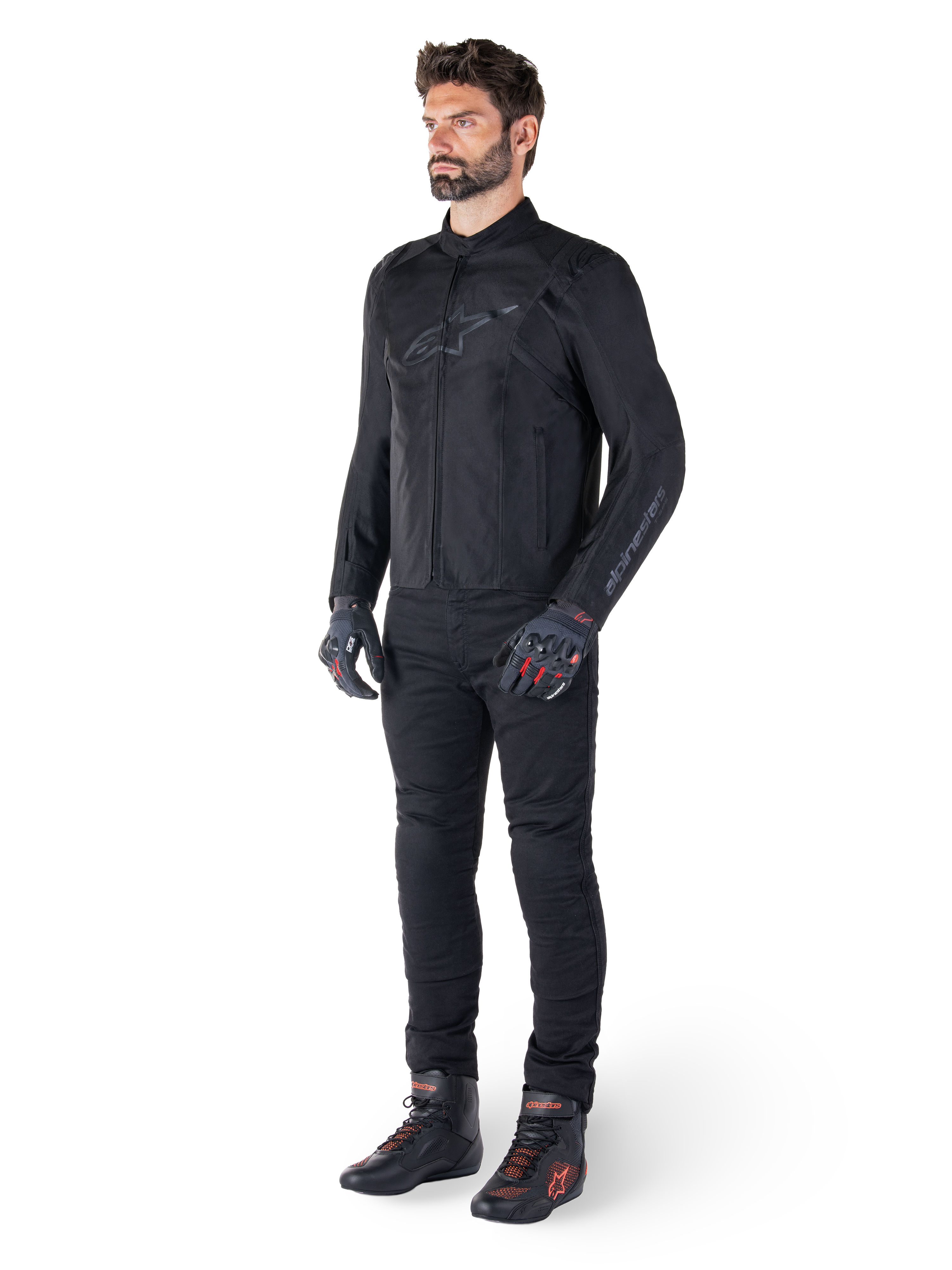 Veste Alpinestars T-SPS V2 Waterproof, noire, portée par un mannequin homme avec un pantalon noir assorti, des gants noirs avec des accents rouges et des chaussures de moto noires, équipement de moto complet pour la conduite urbaine et sportive, fond blanc